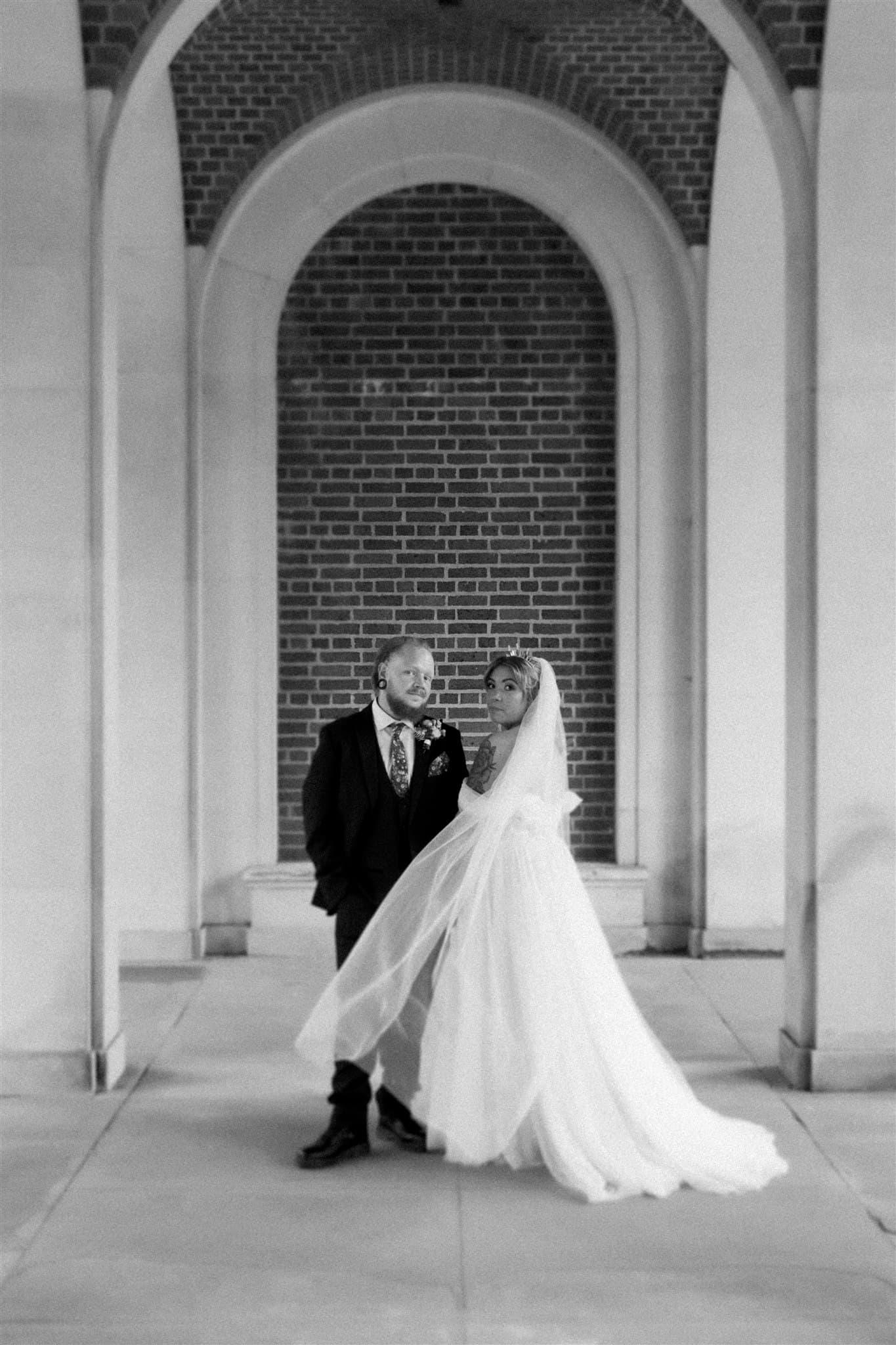 A bride and groom stand under an arched brick doorway. The bride's veil flows gracefully, and both appear serene and elegant in their wedding attire. Photographed by Ivy & Pine