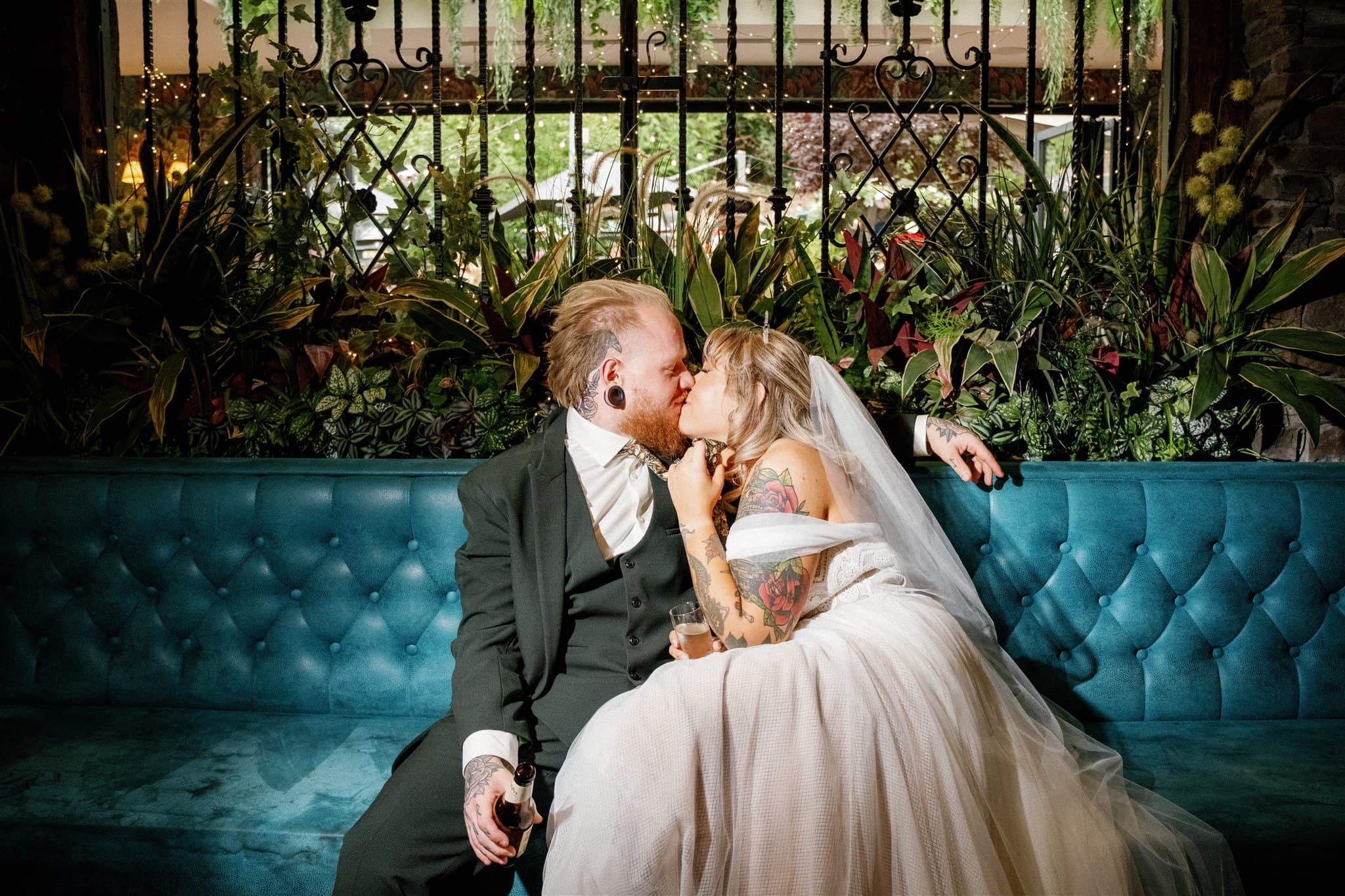 A bride and groom share a kiss on a teal tufted sofa, surrounded by lush plants. The bride wears a white gown, the groom a dark suit, both exuding joy and warmth. Photographed by Ivy & Pine