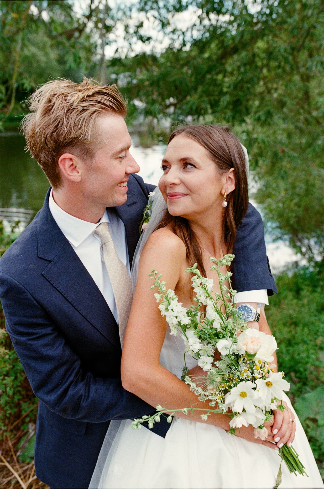 Young couple on their wedding day. The groom in a navy suit embraces the bride in a white dress holding a bouquet, both gazing lovingly at each other with a serene forest background. Photographed on film by Ivy & Pine