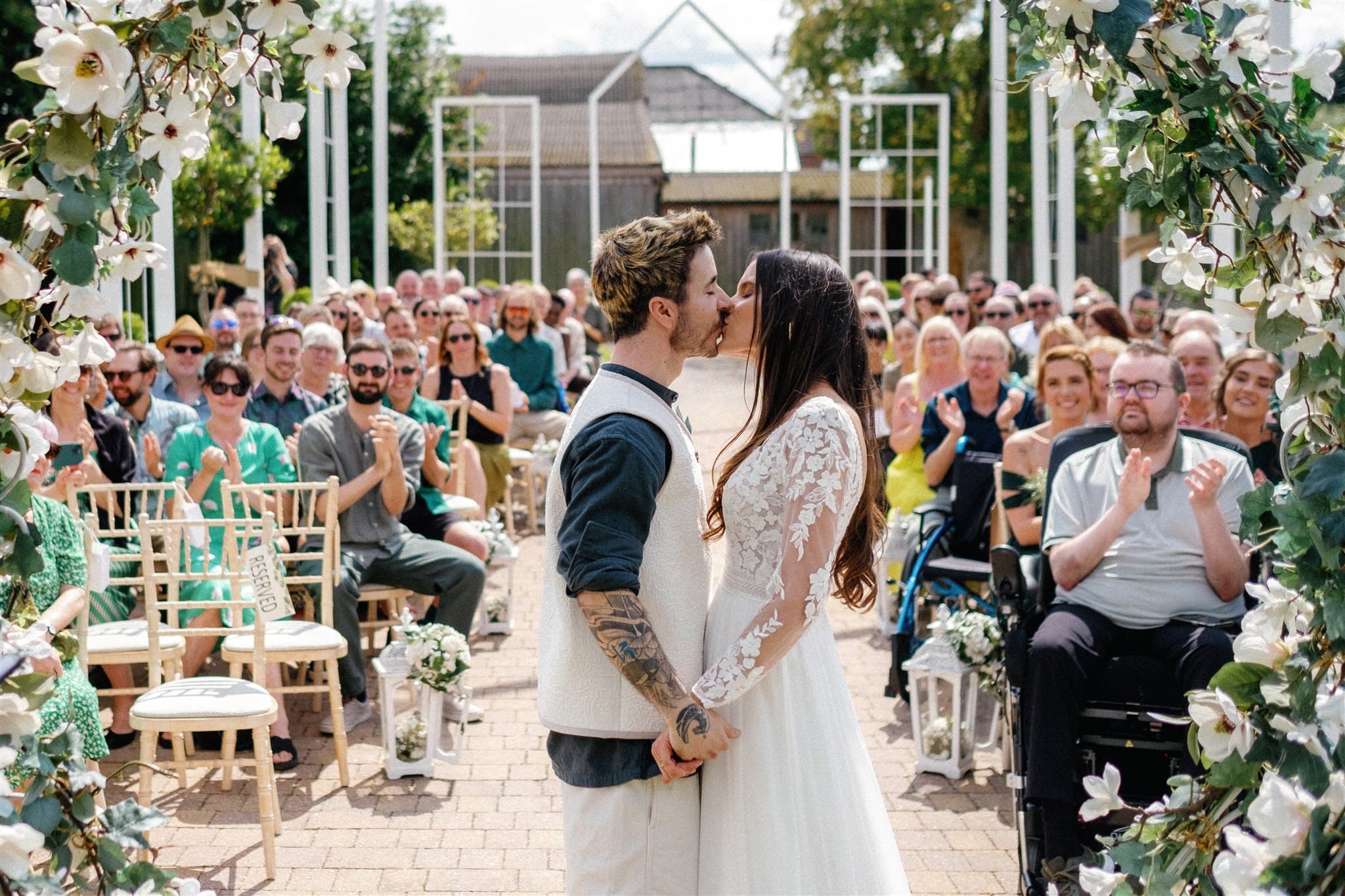 A film photo of a couples first kiss, guests seated on either side, outdoor venue with white flowers.