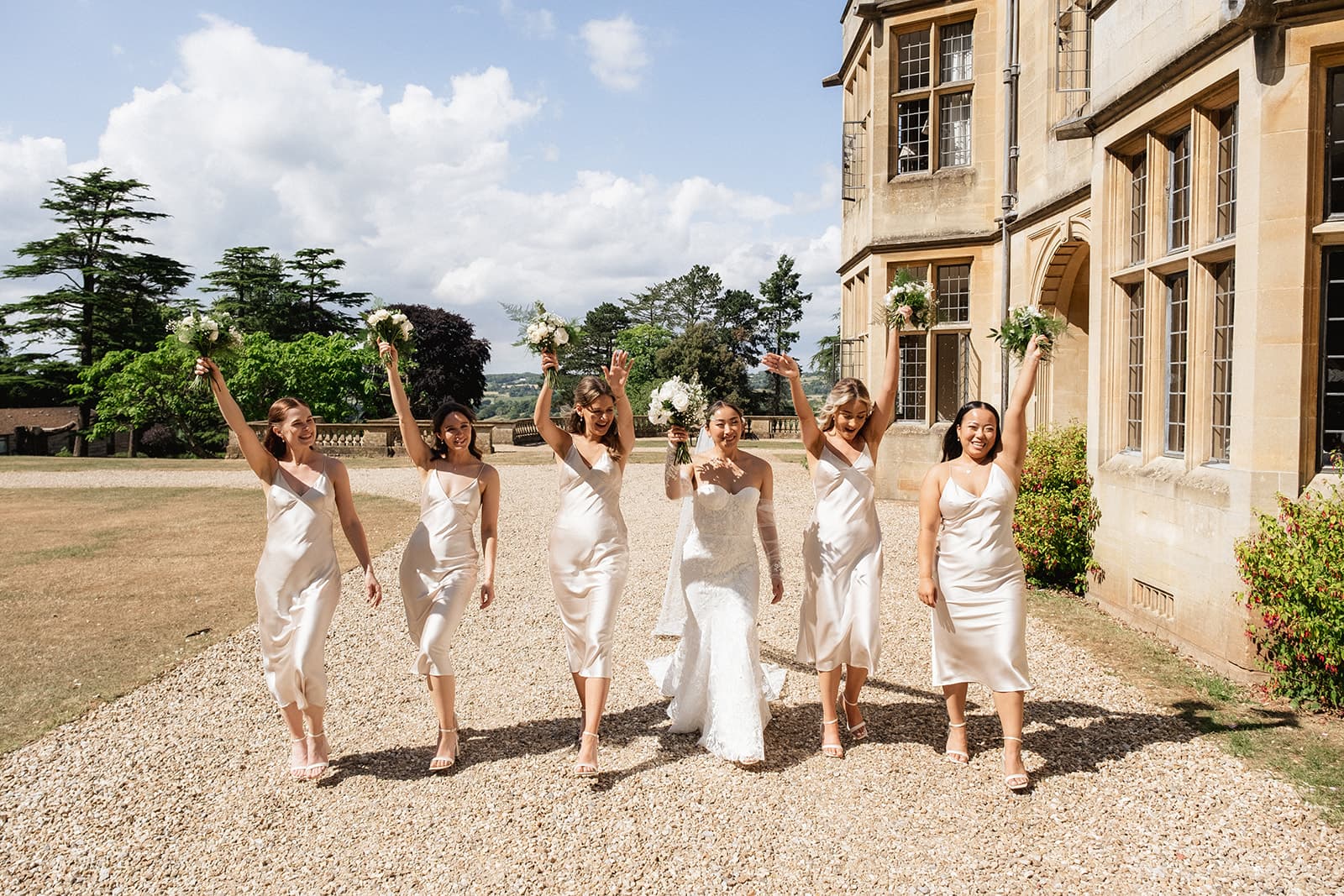 A group of bridesmaids in white dresses holding bouquets next to a stately home.