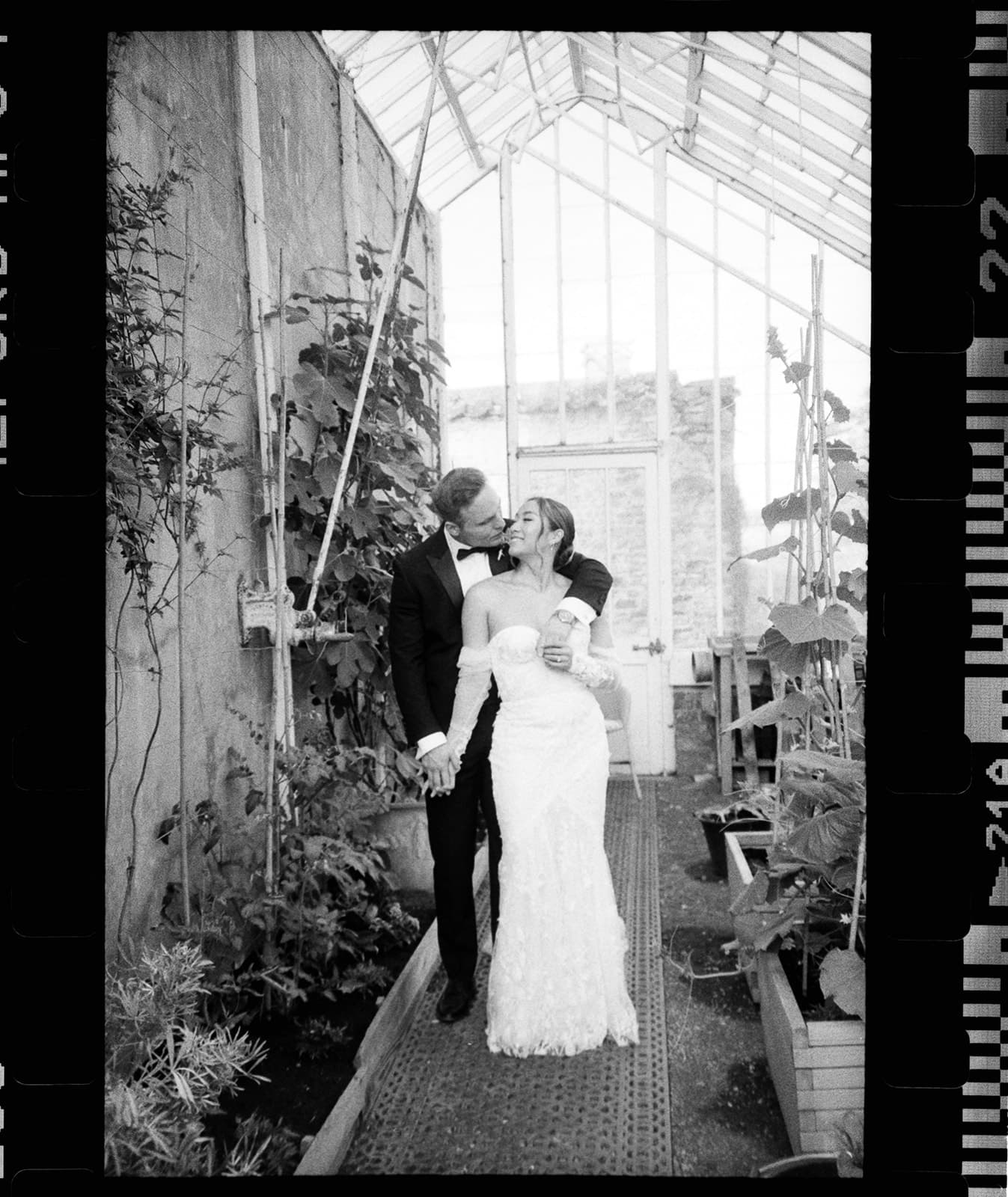 A couple in wedding attire embracing in a sunlit greenhouse.