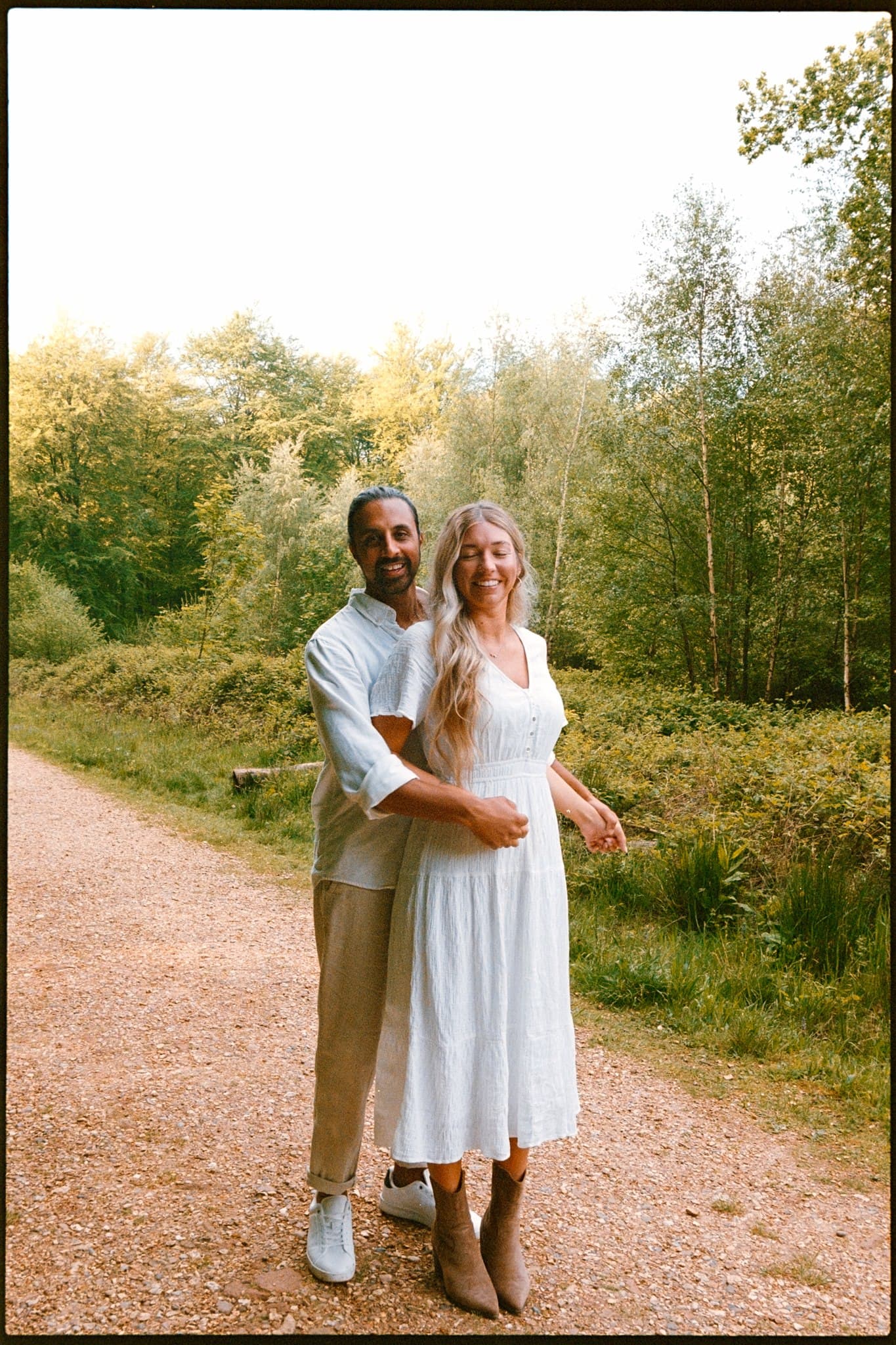 A couple standing close together on a forest path, woman in white dress, man in light shirt and trousers.