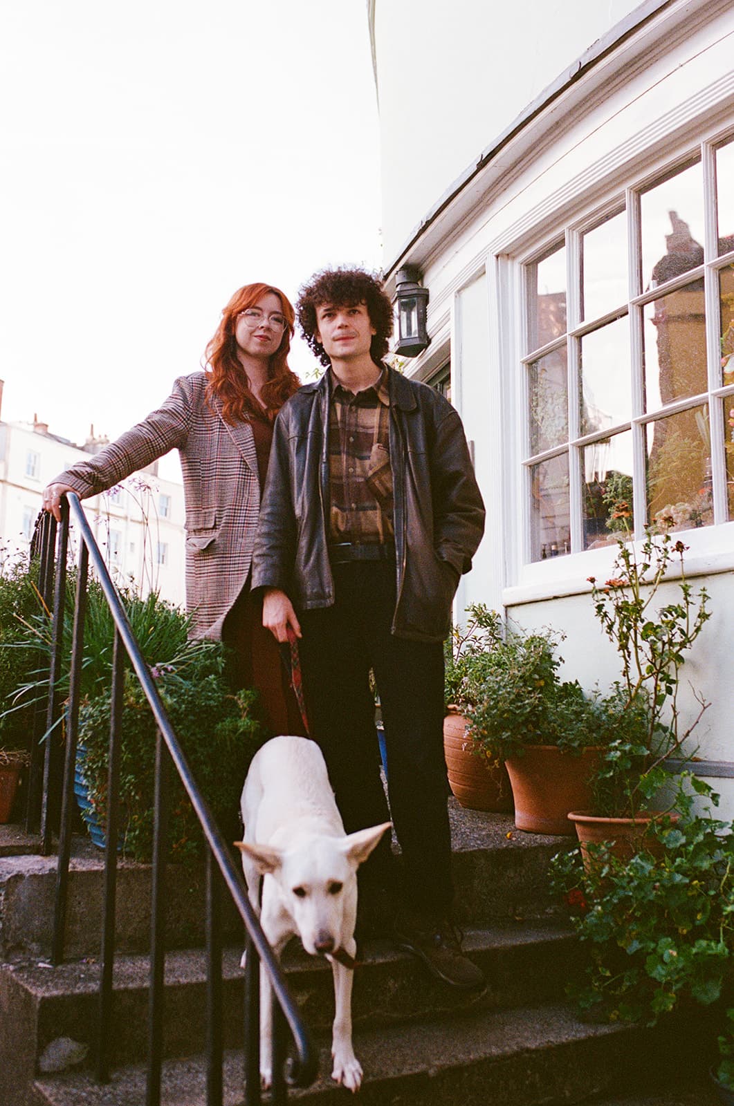 Two people with a dog on house steps surrounded by potted plants.