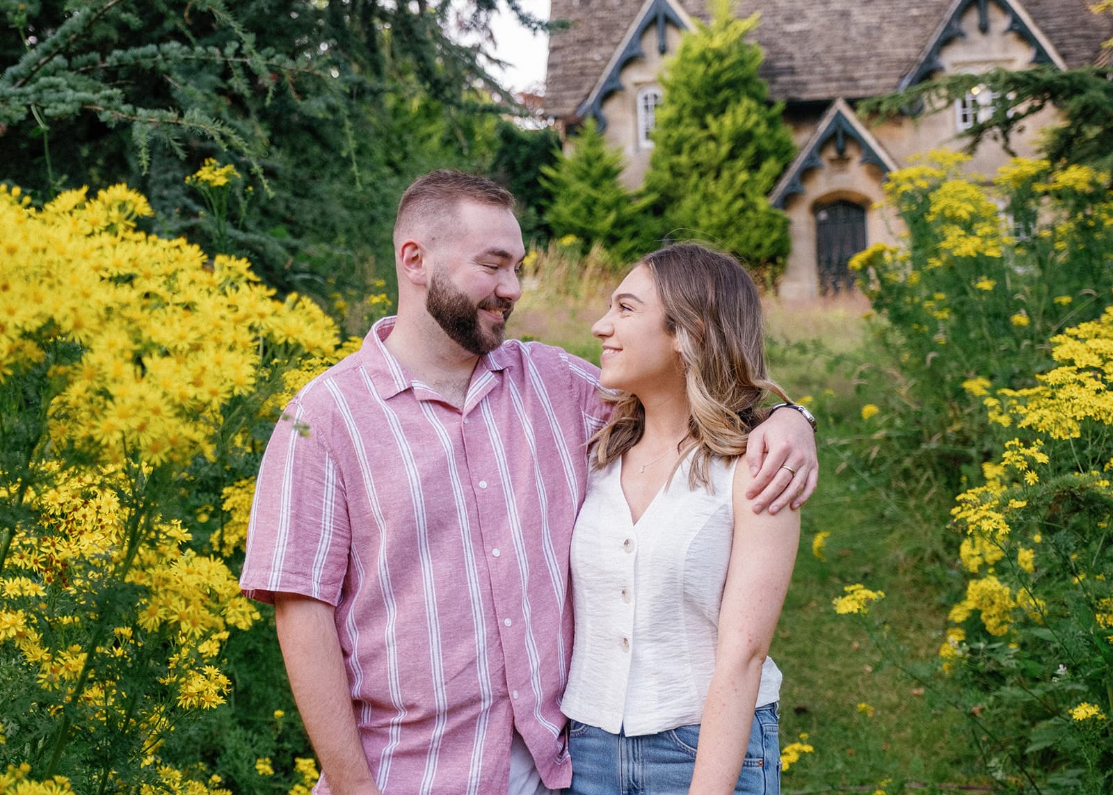 A couple standing in front of yellow flowers with a house in the background.