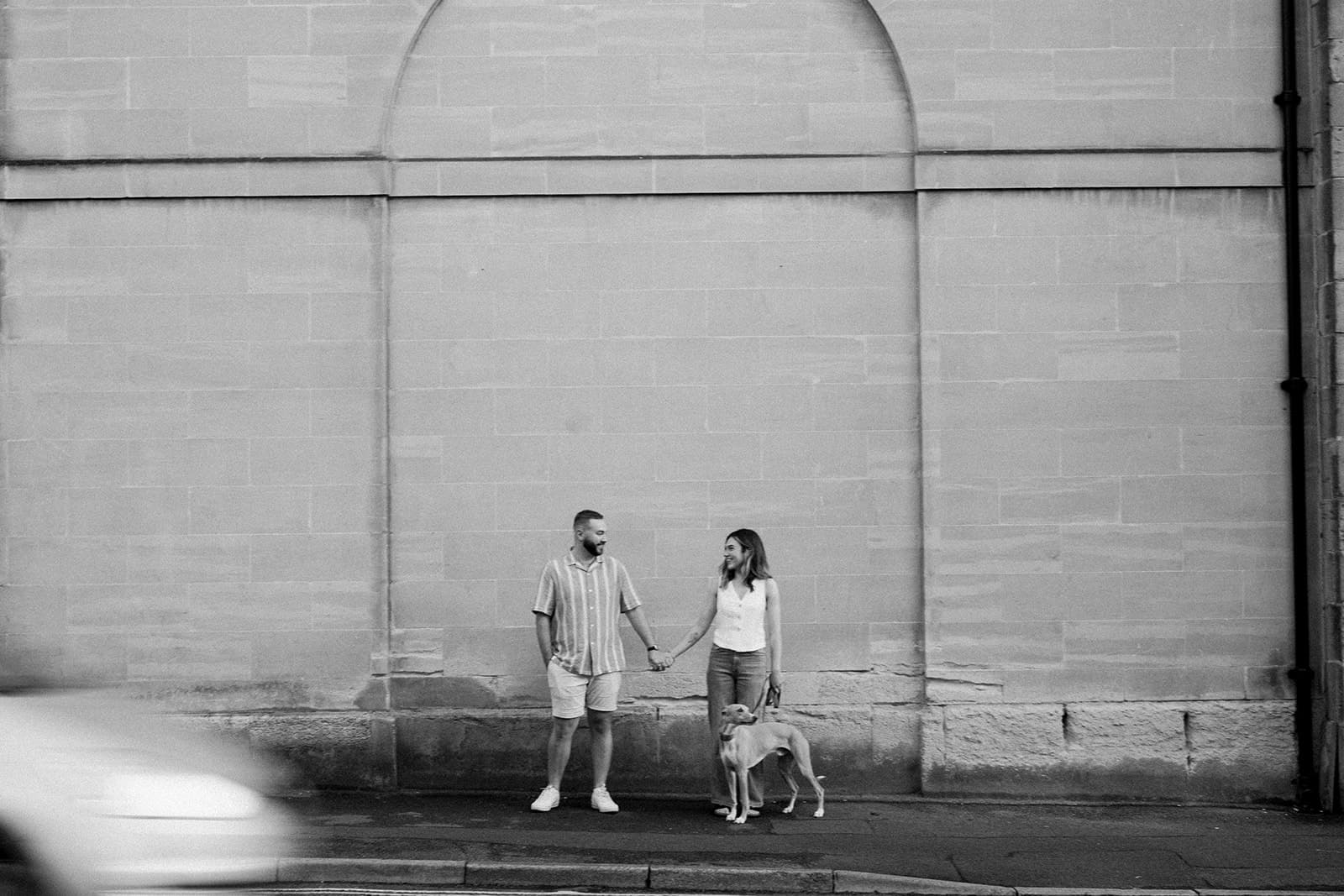 Two people holding hands with a dog, standing by a wall.