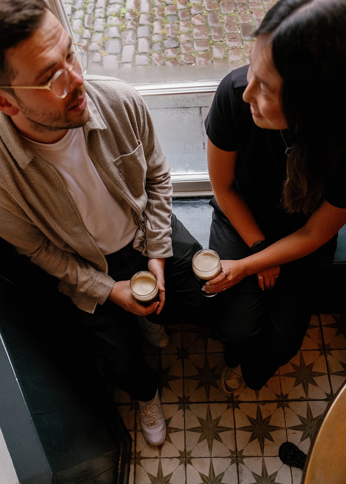 Two people holding Gunniess, sitting by a window in the Ostrich, Bristol, with patterned floor tiles.