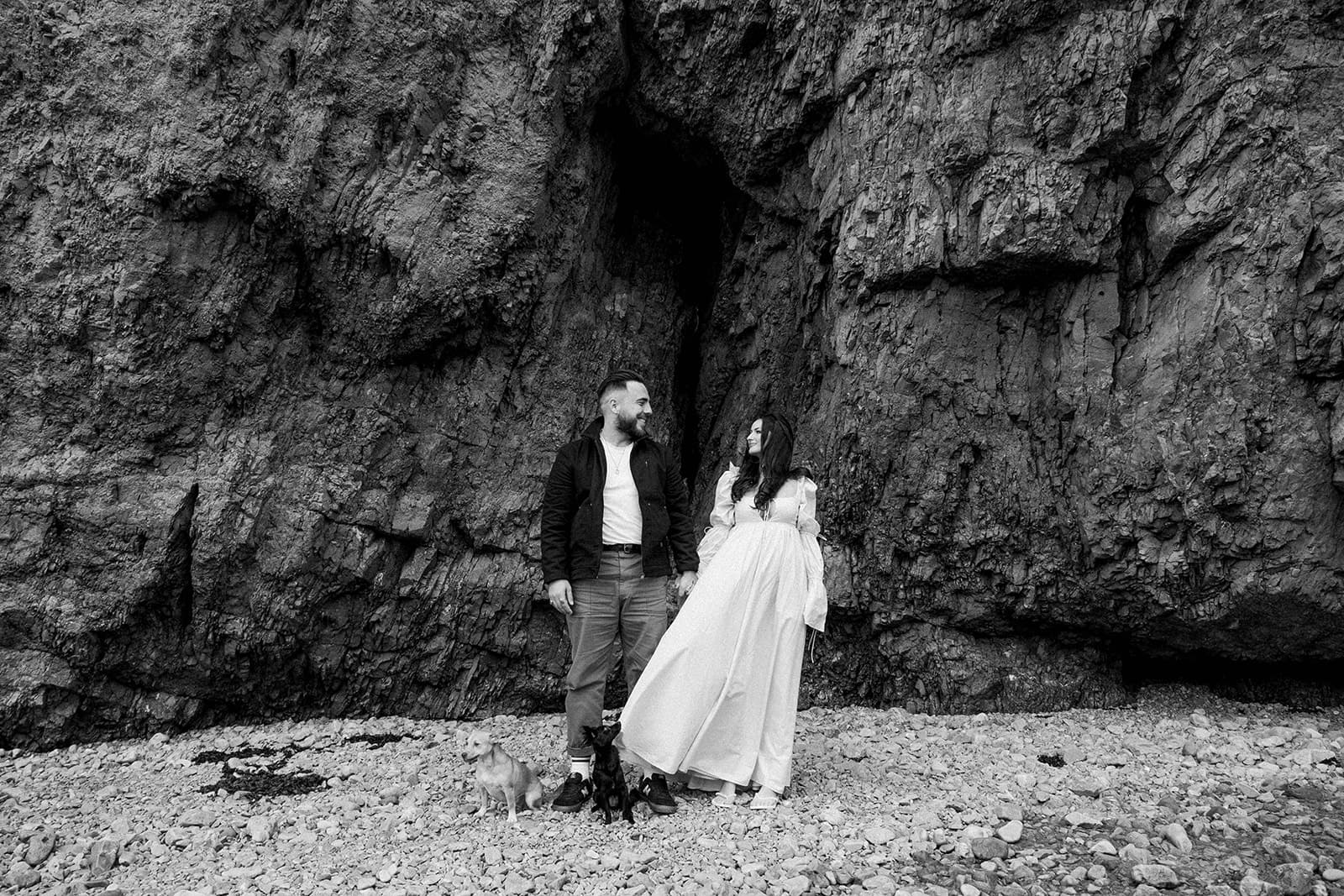 A couple with dogs standing by a rocky cave entrance.