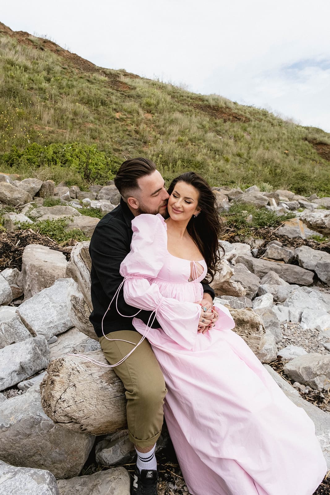 Couple sitting closely on rocky terrain with grassy hill in the background.