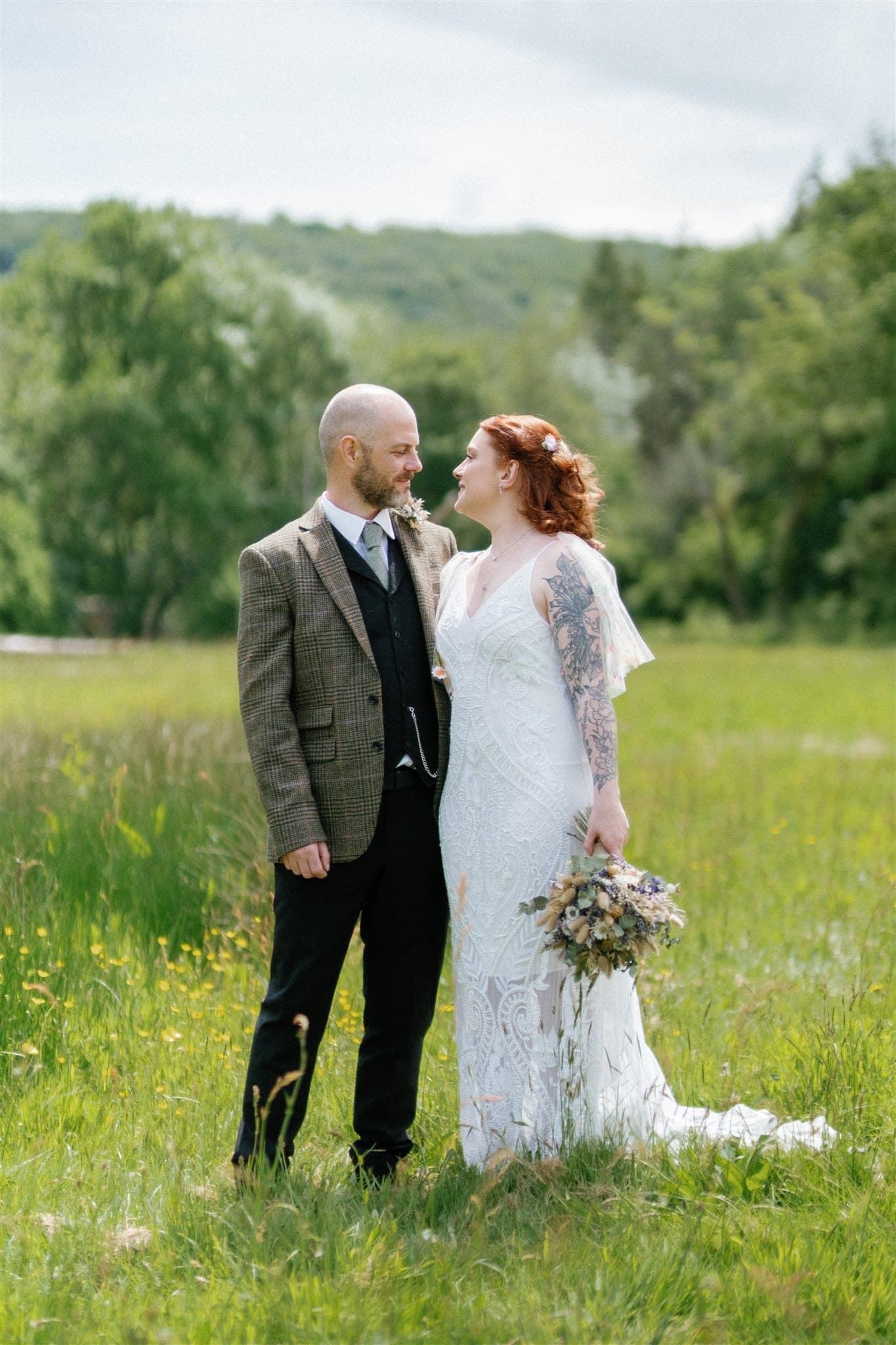A couple in wedding attire standing in a green field with trees in the background.