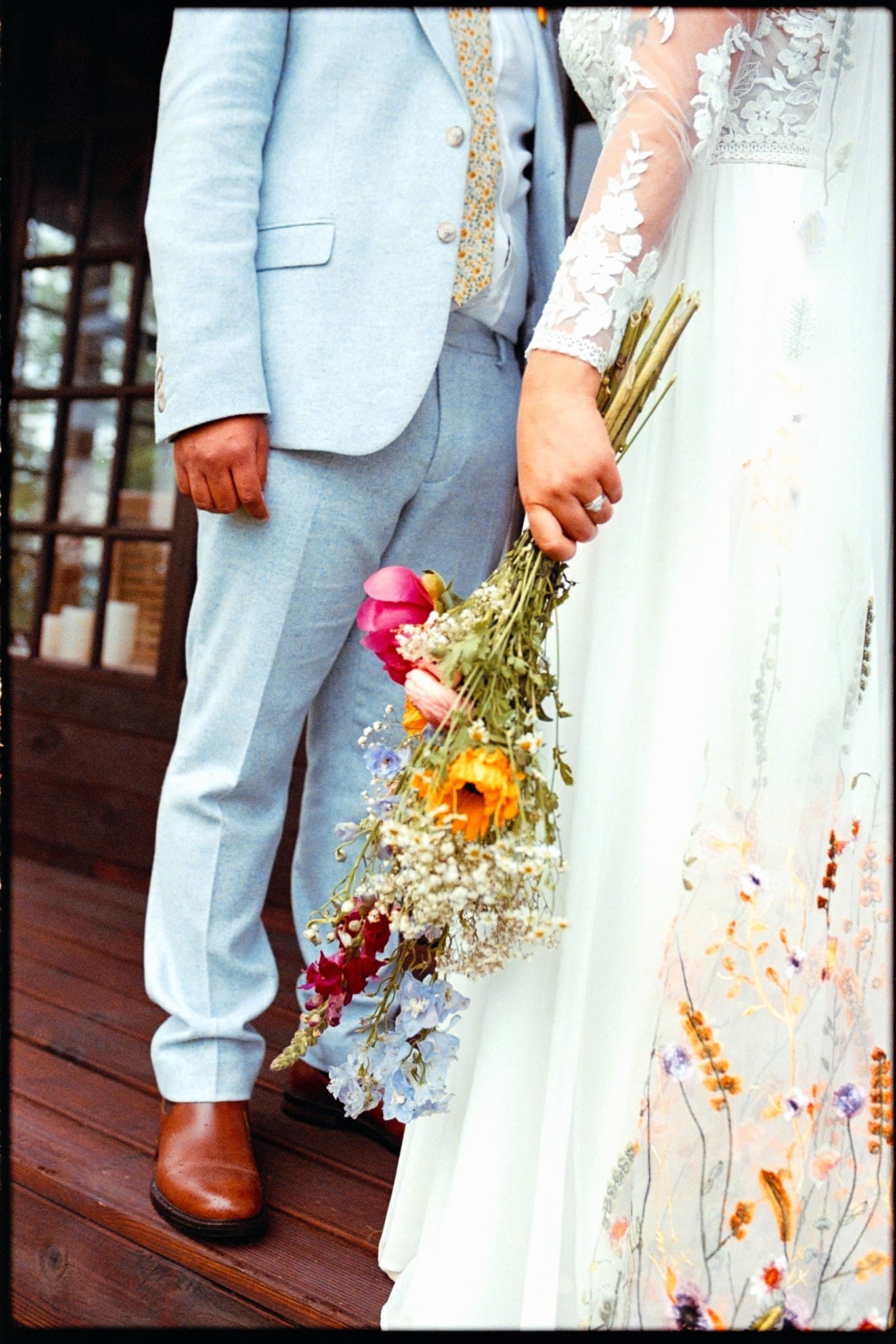 Bride and groom holding hands, partial view, with a vibrant flower bouquet. Film Photo