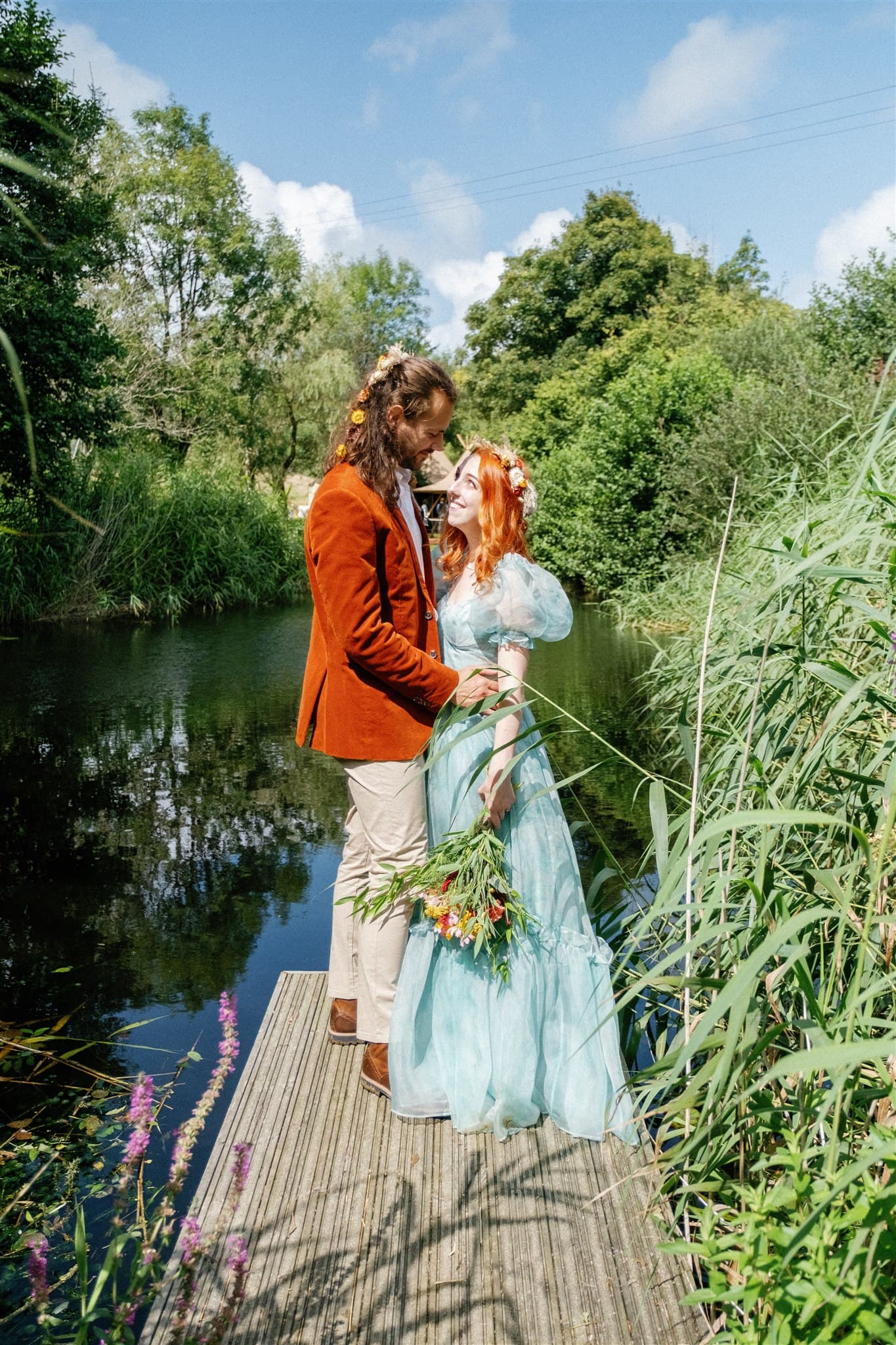 A couple in wedding attire standing on a jetty surrounded by greenery.