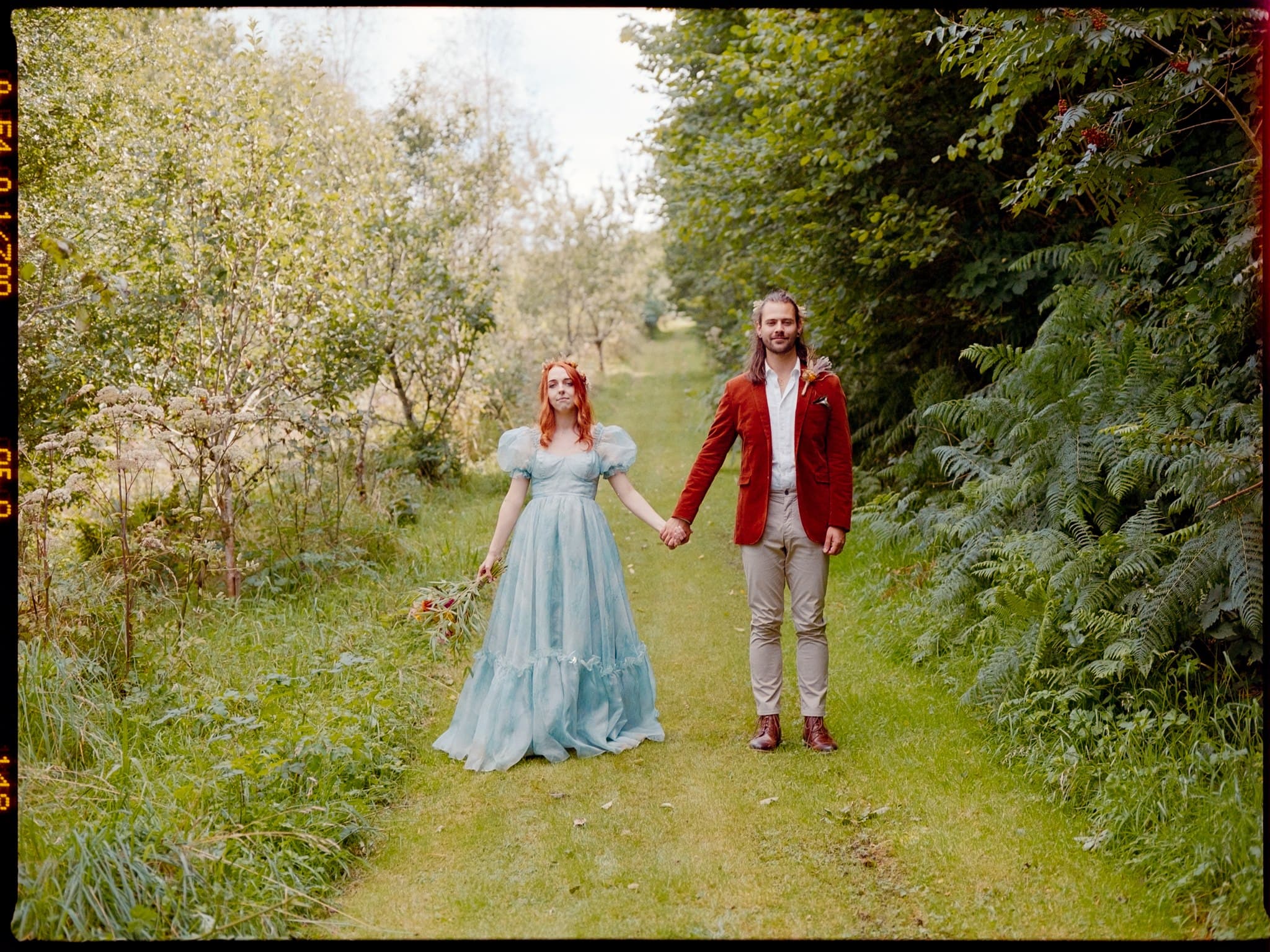 A film photo of a wedding couple holding hands in a garden path, one in a blue wedding dress and the other in a red jacket.