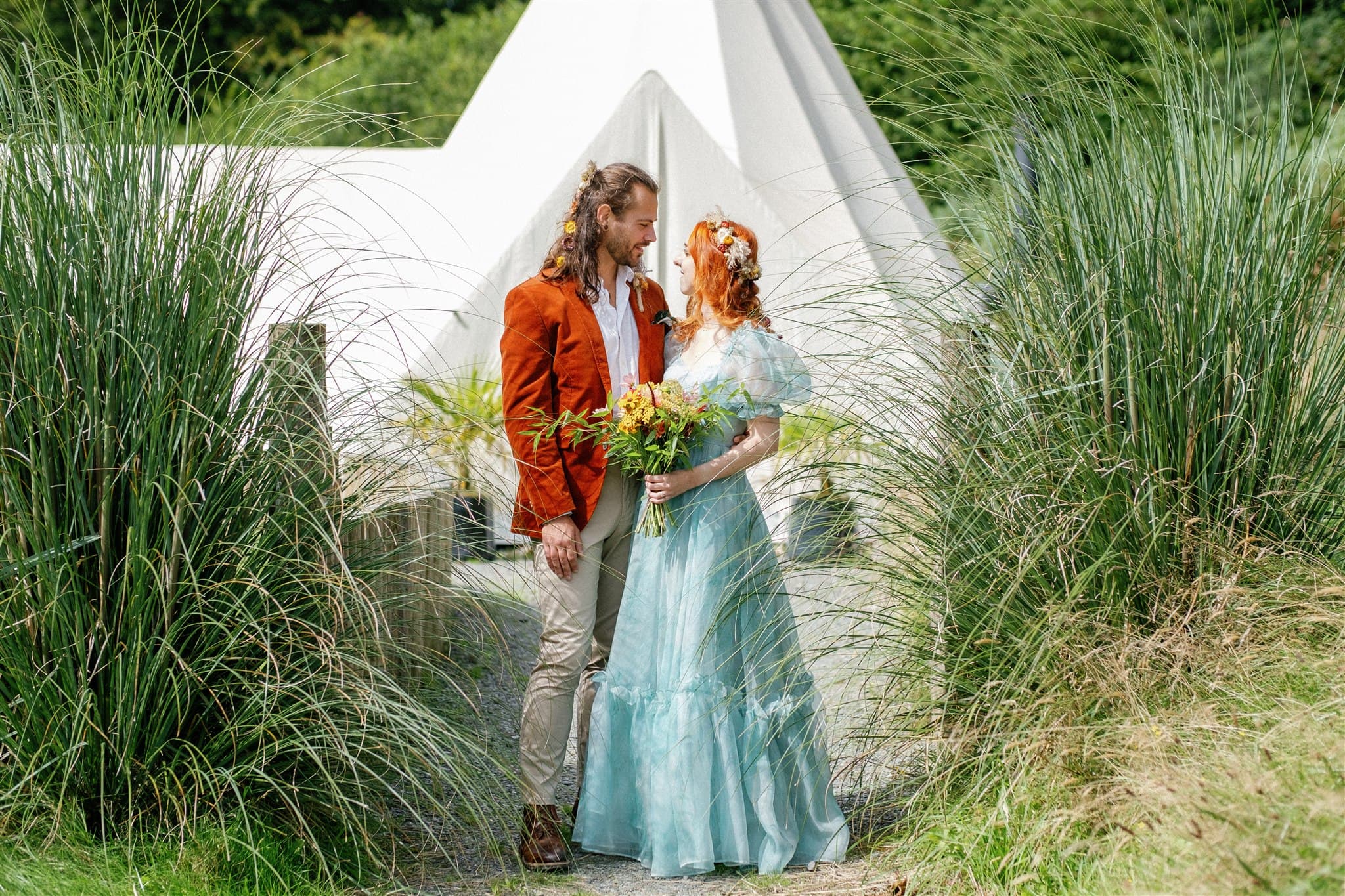 Couple in wedding clothes holding hands in front of a tipi, surrounded by tall grass.
