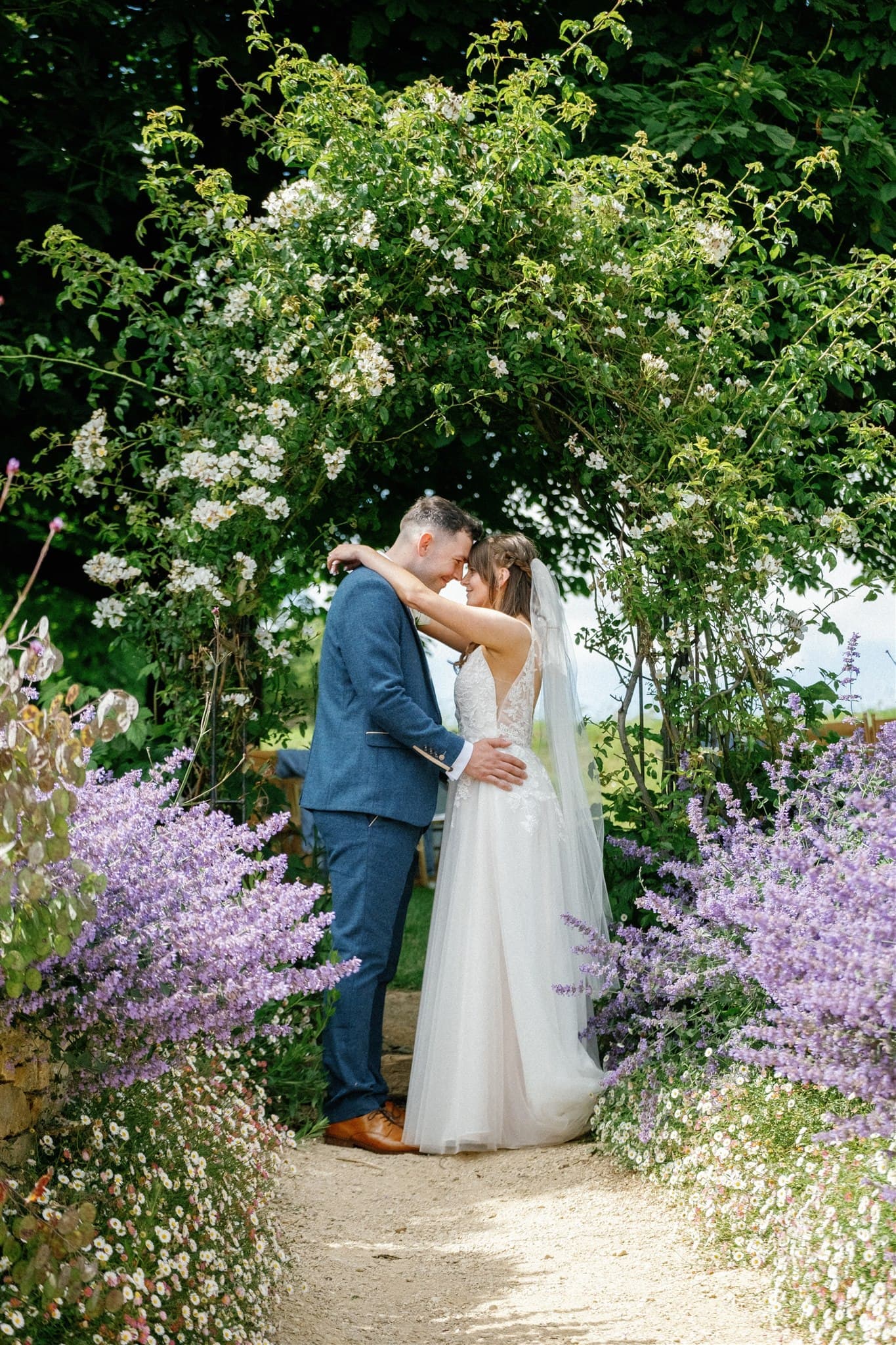 A couple in wedding attire embracing under an arch of white flowers and greenery.