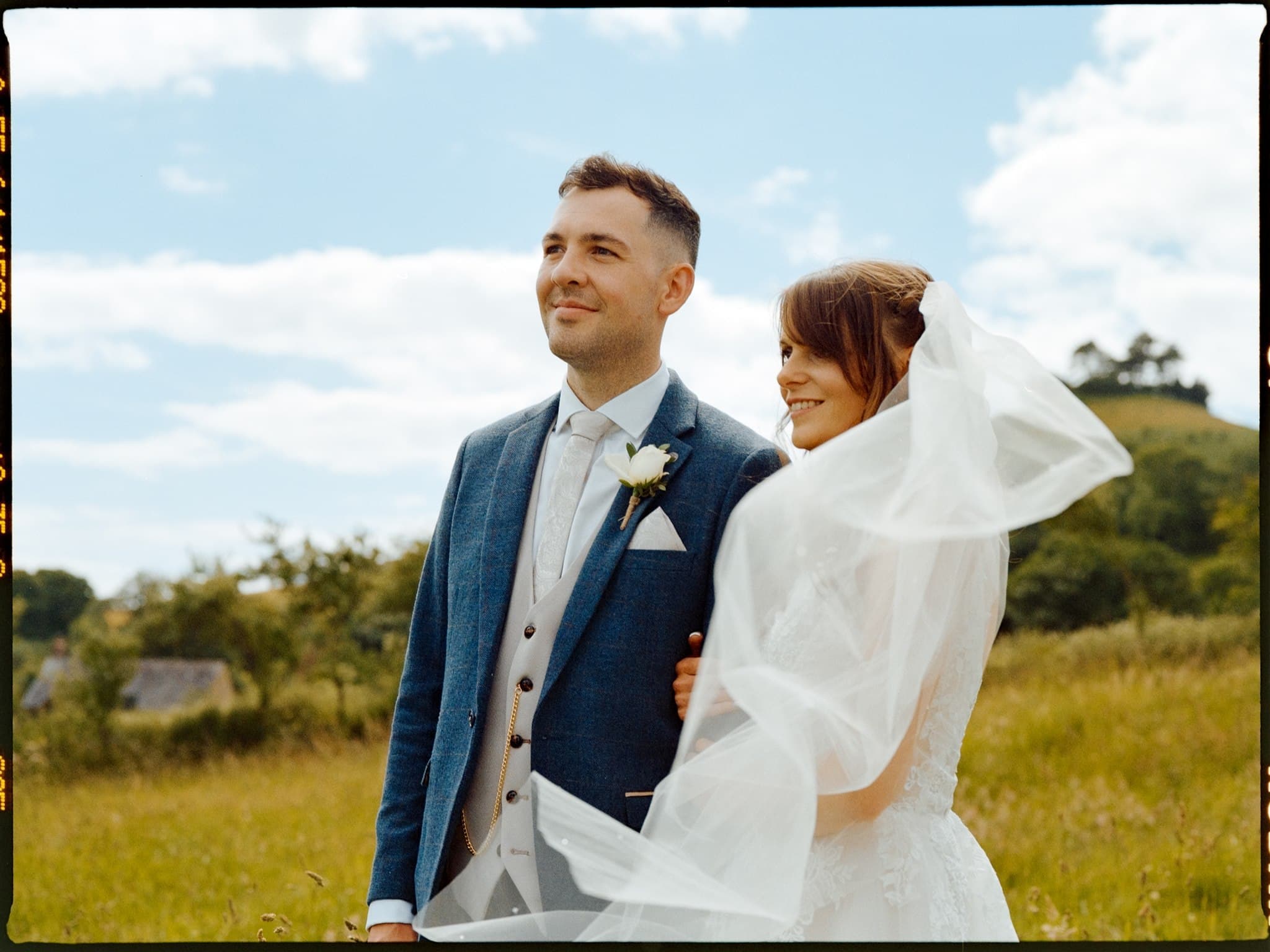 Film photo of Wedding couple standing in a field, groom in a blue suit, bride with a veil.