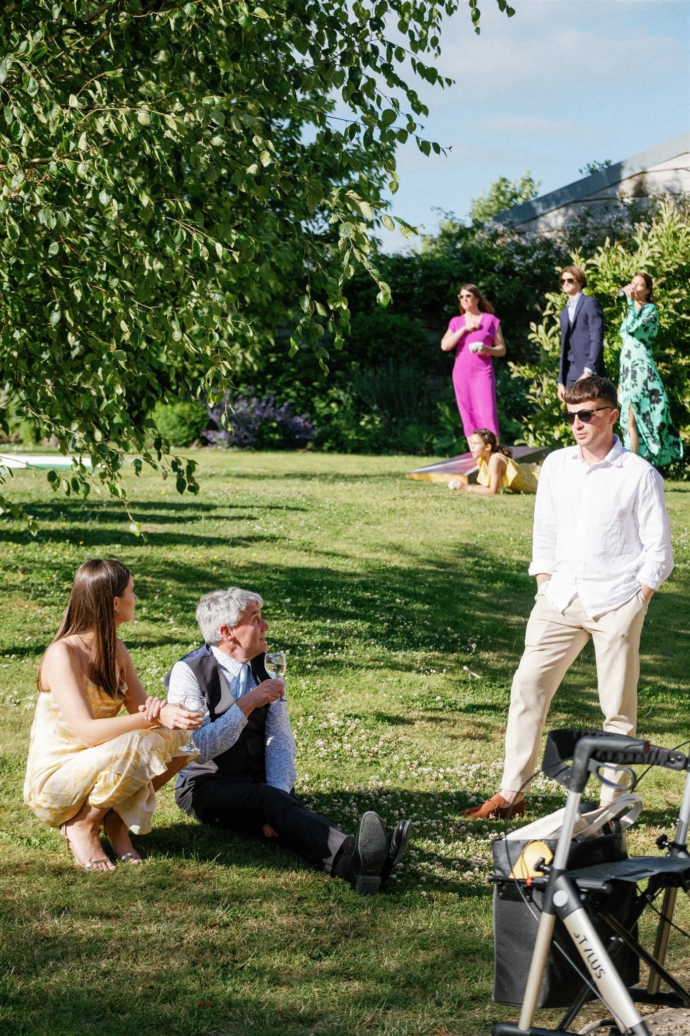 Film photo of People in elegant attire enjoying a sunny day in a green field with trees and a bicycle in the foreground.