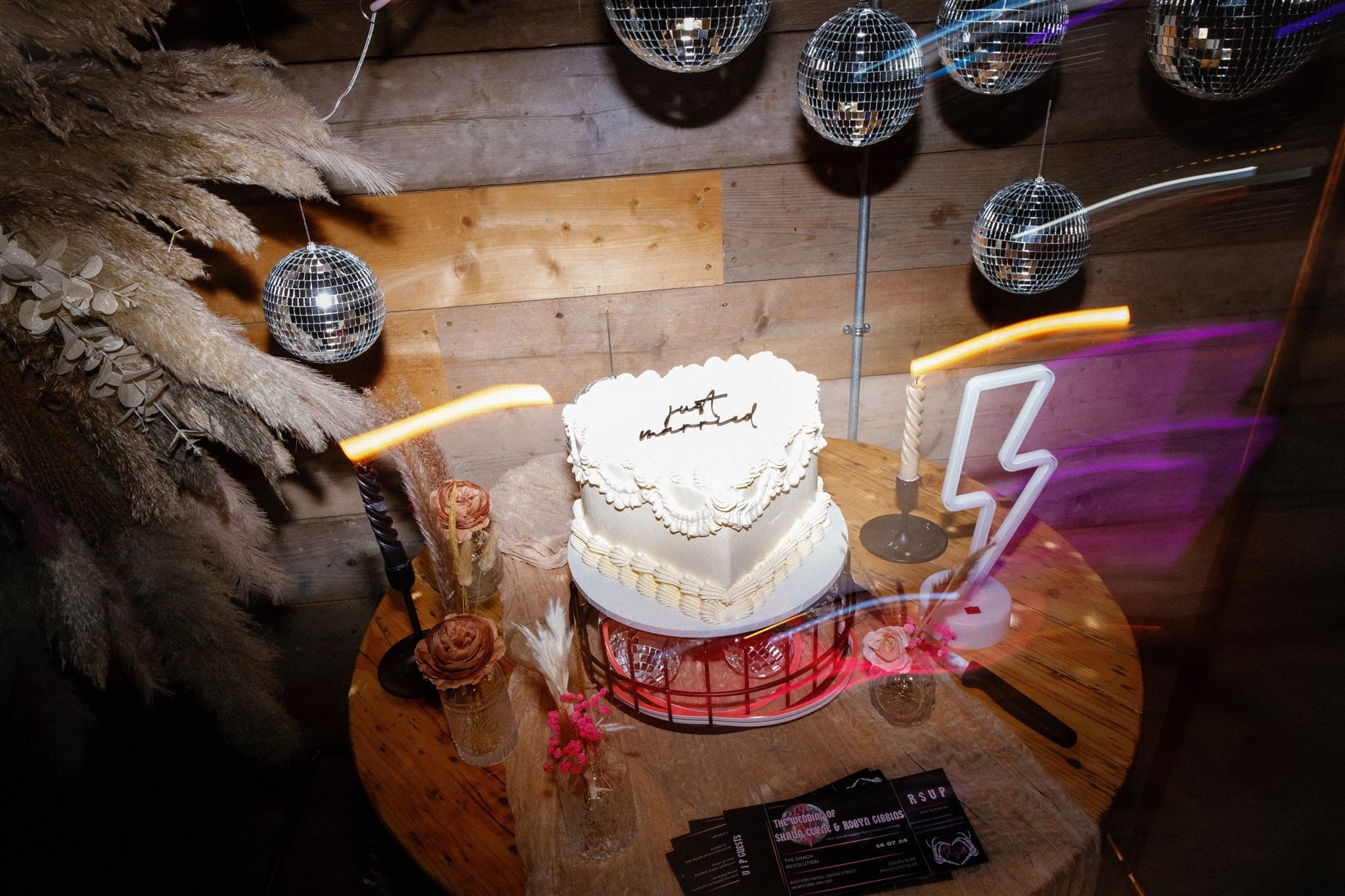 Tiered white cake with "just married" on top, surrounded by disco balls, flowers, and neon light.