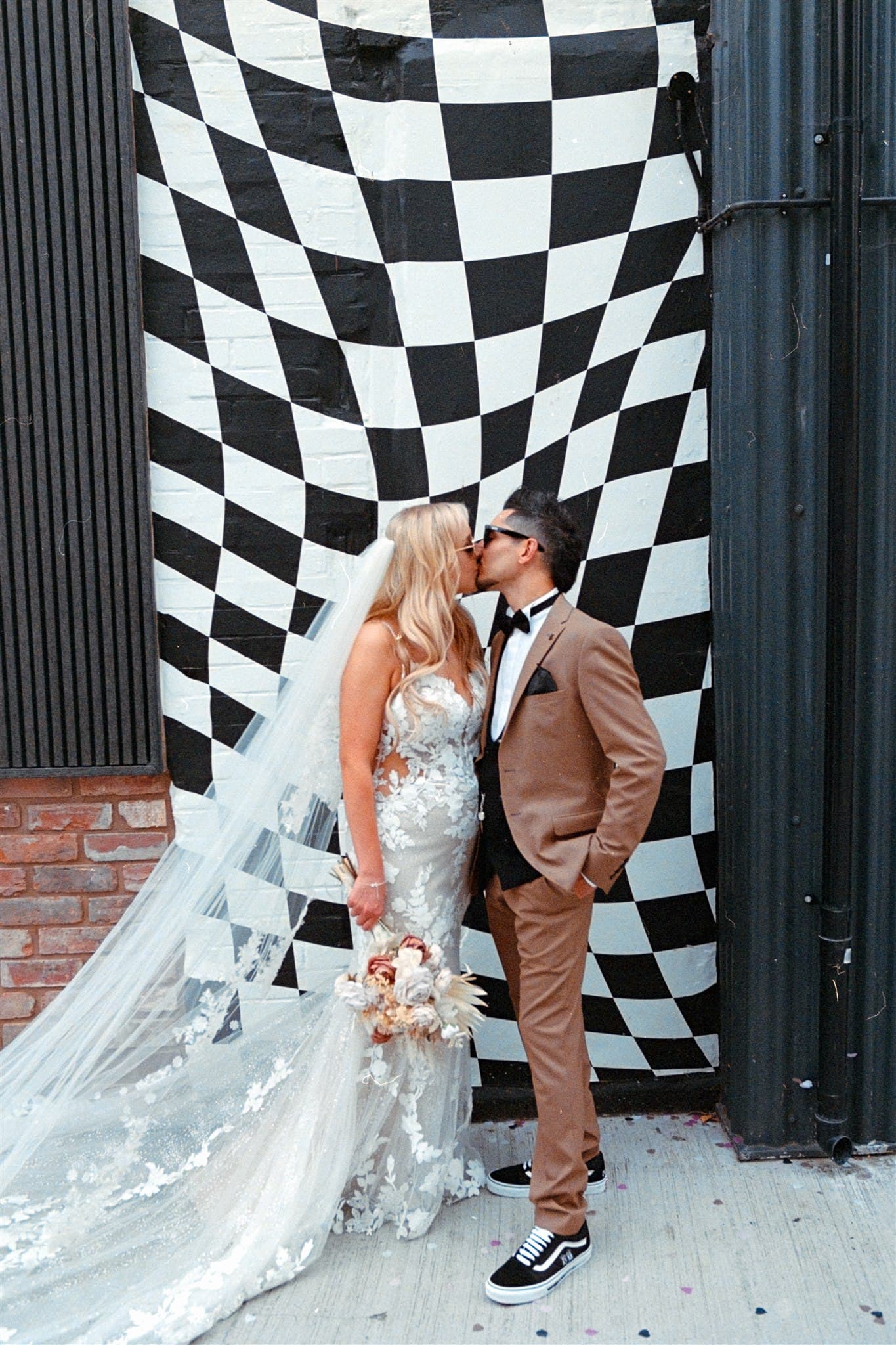 A couple in wedding attire standing in front of a black-and-white checkered wall.