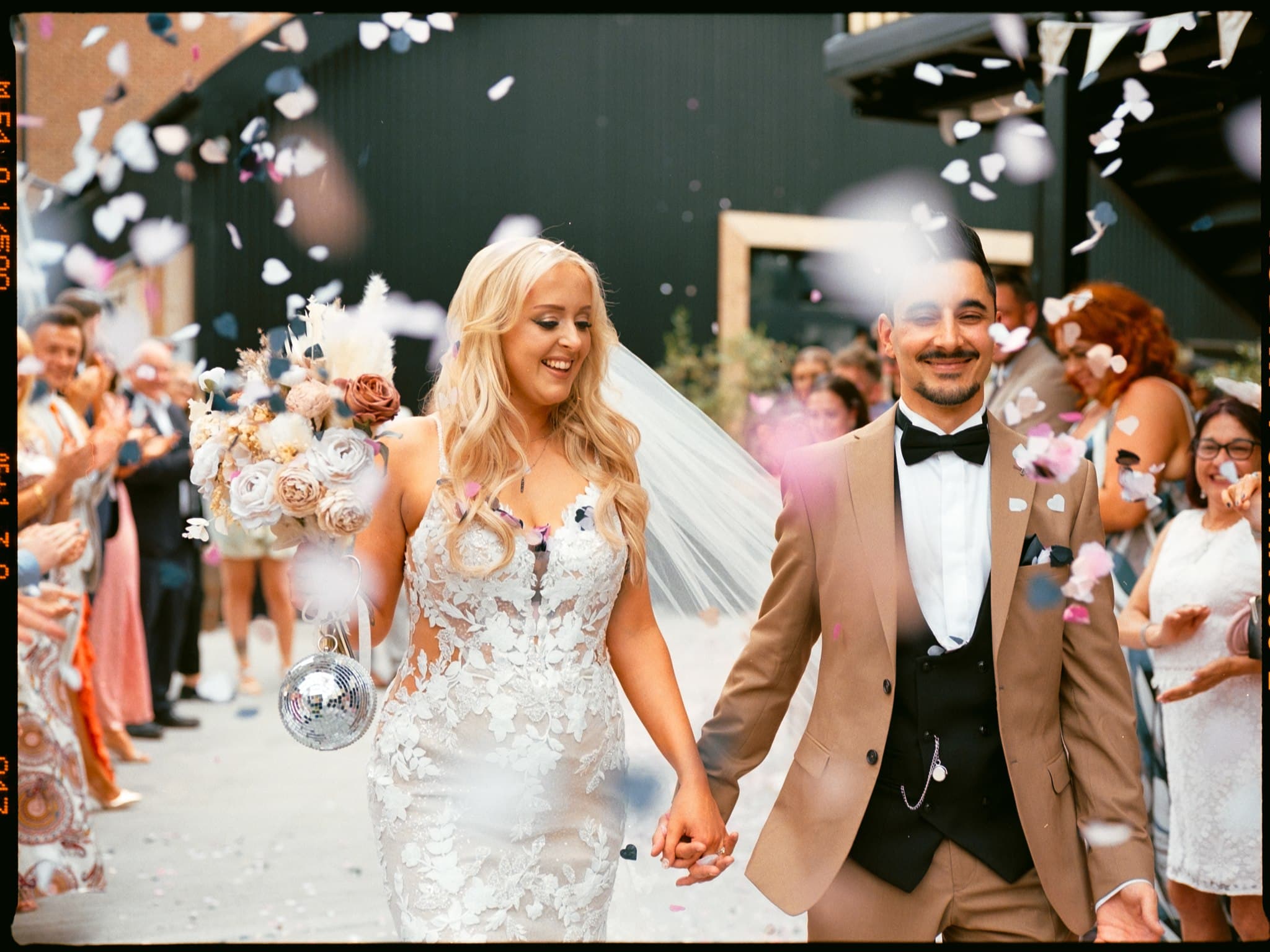 A film photo of wedding couple walking through confetti, guests in colourful attire cheering.