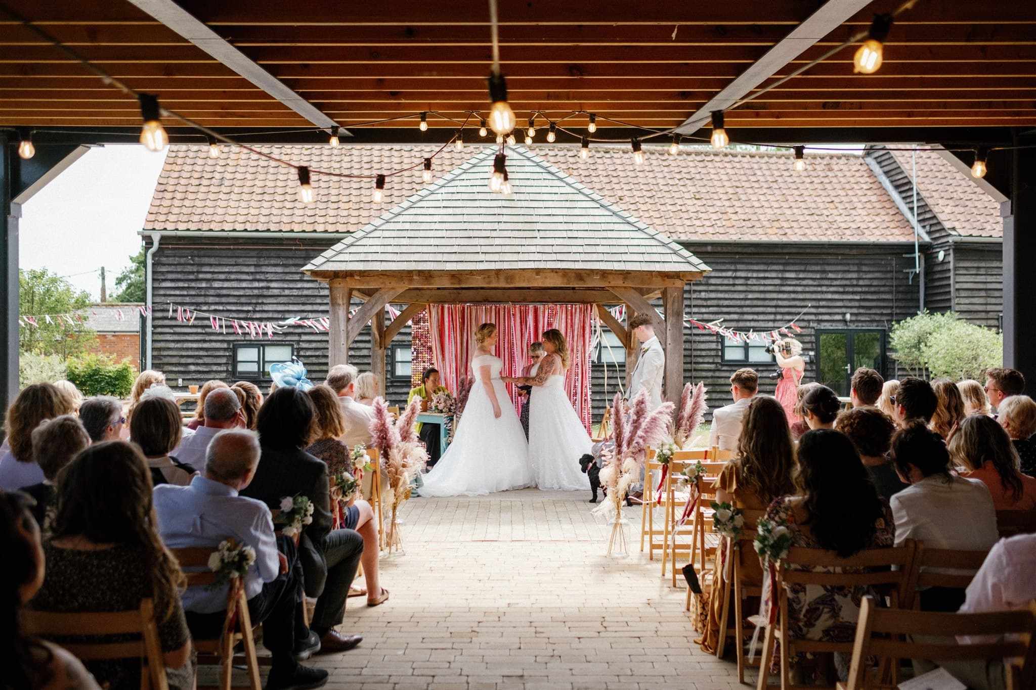 A joyful wedding scene with two brides in white dresses under a gazebo, surrounded by seated guests. The venue is rustic with string lights and festive decor. Photographed by Ivy & Pine