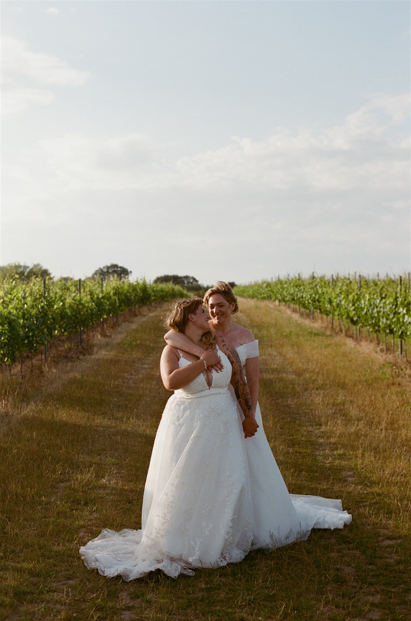 Two brides in white wedding dresses embrace and smile in a sunlit vineyard, conveying joy and love. The sky is clear with a few clouds. Photographed by Ivy & Pine