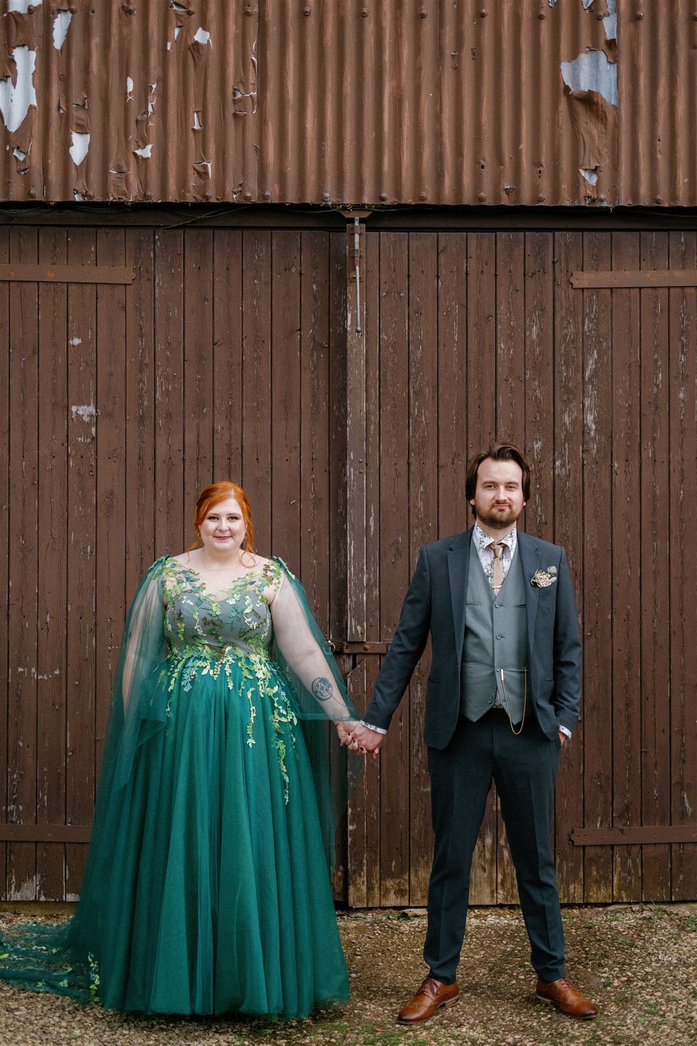 A couple in wedding outfits holding hands in front of a rustic wooden door and corrugated metal wall.