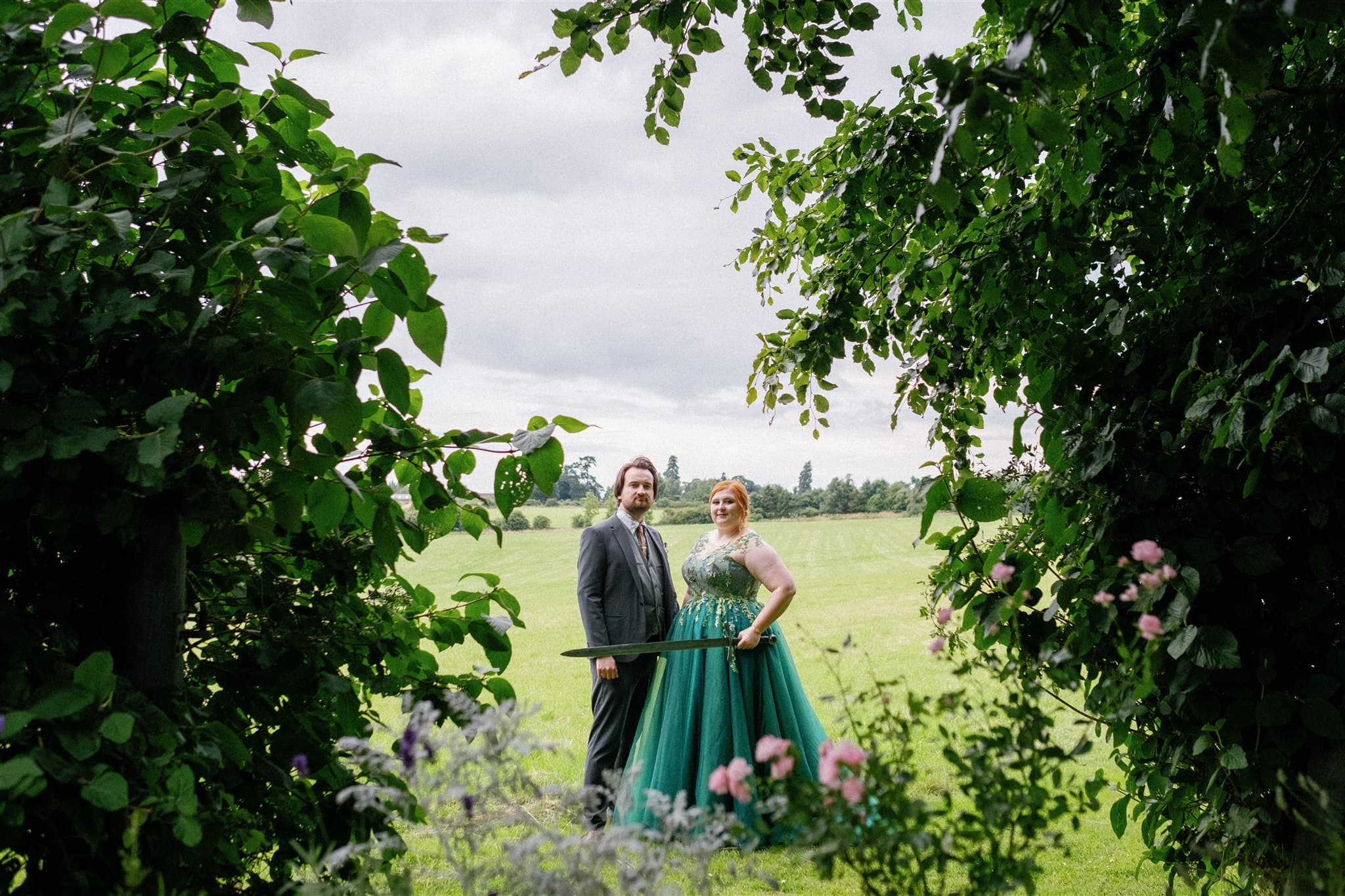 Bride and Groom standing in a lush green field framed by leaves holding a sword at a fantasy themed wedding.