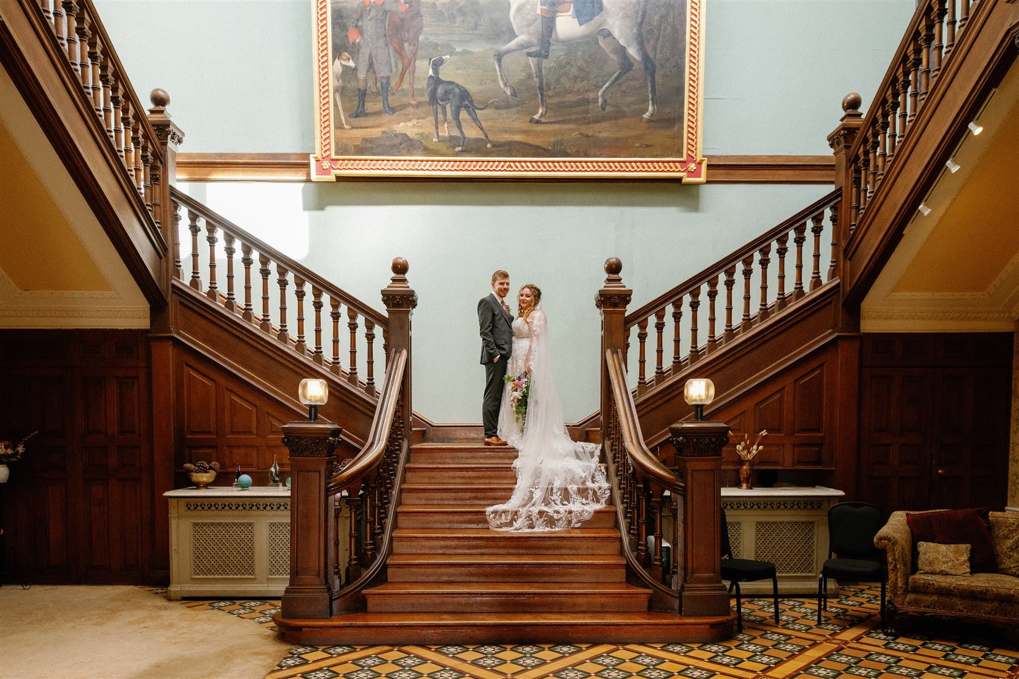 A bride and groom stand on an ornate wooden staircase. The bride wears a white gown with a long train, holding flowers. A large painting hangs above them, adding elegance. Photographed by Ivy & Pine