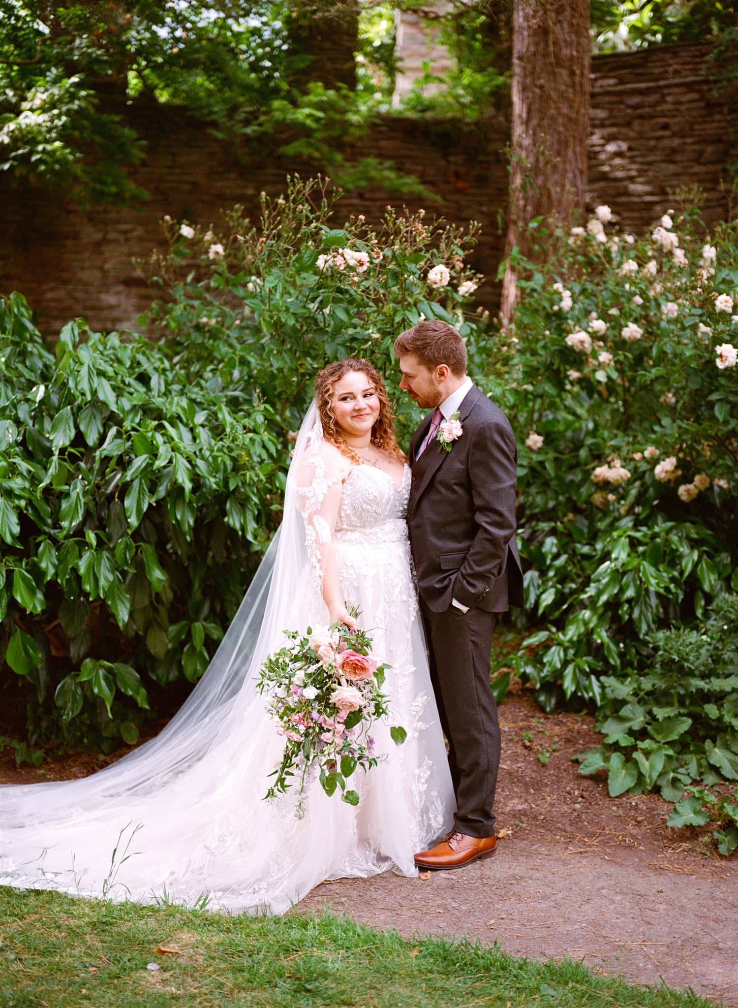 Bride and groom stand embraced in a garden, surrounded by lush greenery and blooms. The bride holds a bouquet, wearing a flowing lace dress. Romantic and serene. Photographed on film by Ivy & Pine