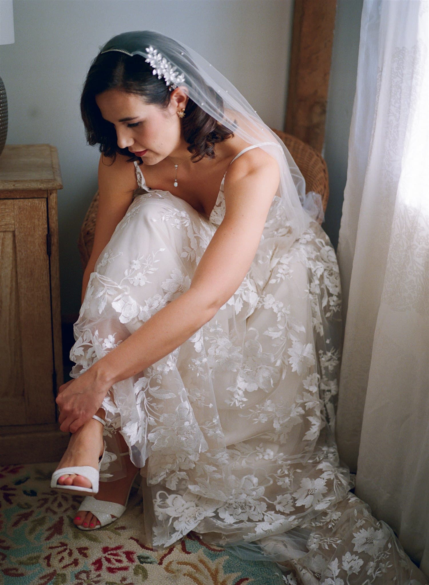 A film photo of Bride in a floral wedding dress putting on her shoe by a window.