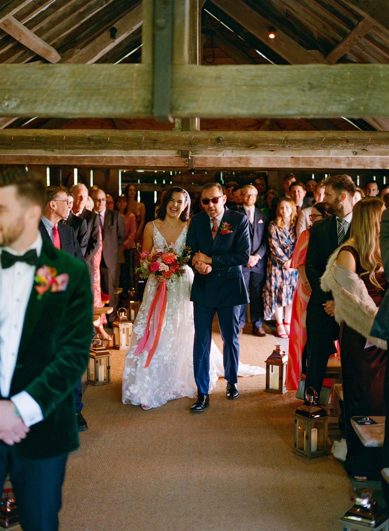 A film photo of A bride and groom holding hands at a rustic wedding aisle, surrounded by guests and lanterns.