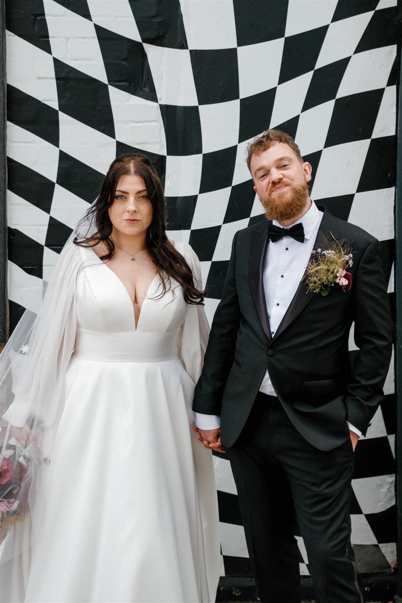 A bride and groom pose before a checkered backdrop. The bride in a white gown holds the groom's hand, while he wears a black suit with a floral boutonniere. They both appear calm and composed. Photographed by Ivy & Pine