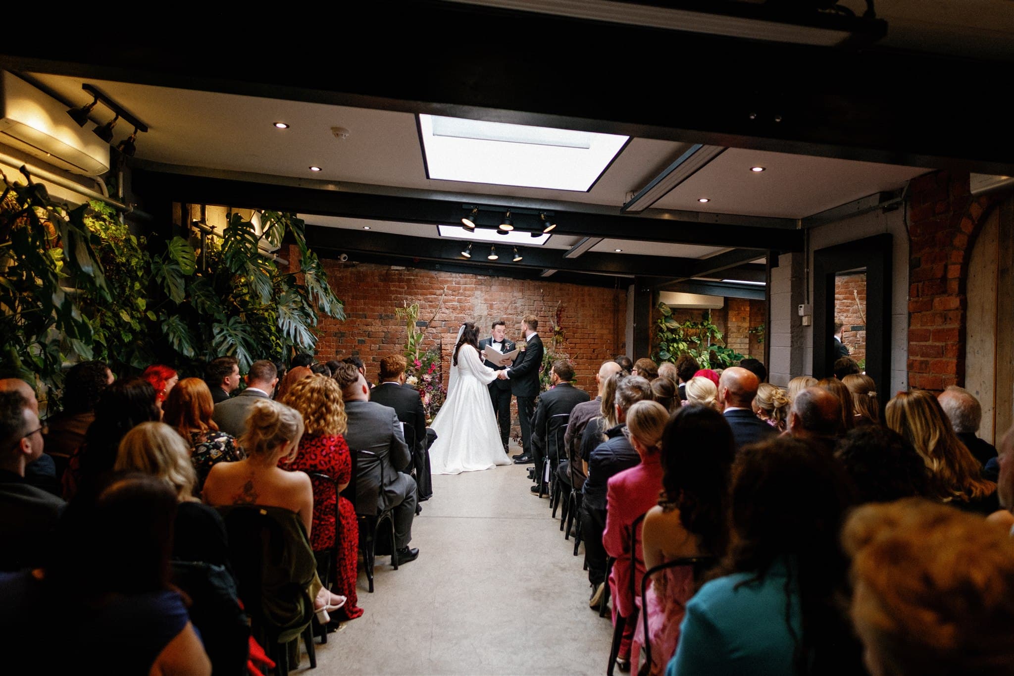 A bride and groom exchange vows in an intimate indoor wedding with exposed brick walls and lush greenery. Guests sit closely, creating a warm, joyful atmosphere. Photographed by Ivy & Pine