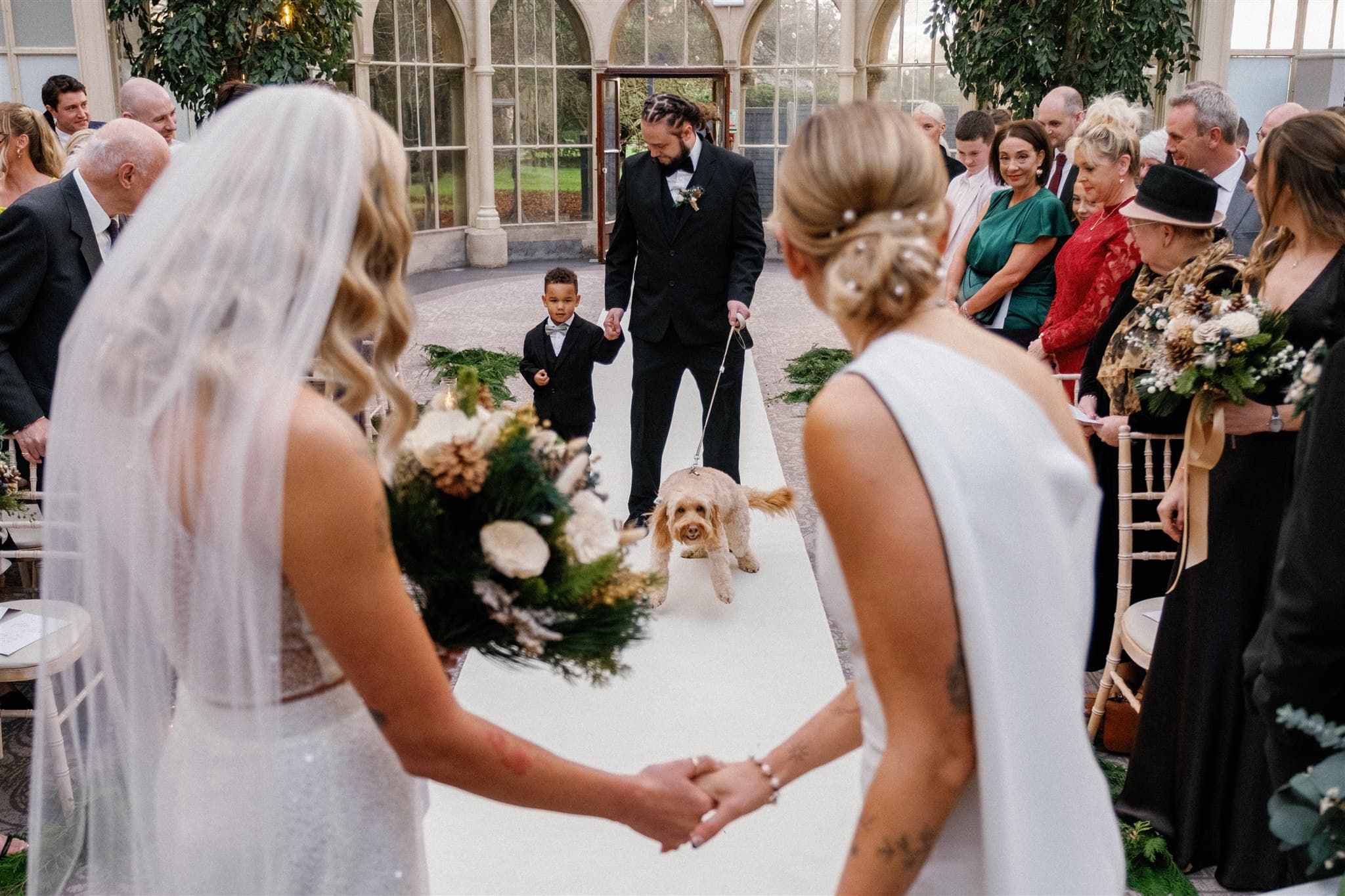 Wedding scene with a bride and bride holding hands, surrounded by guests and a dog leading.