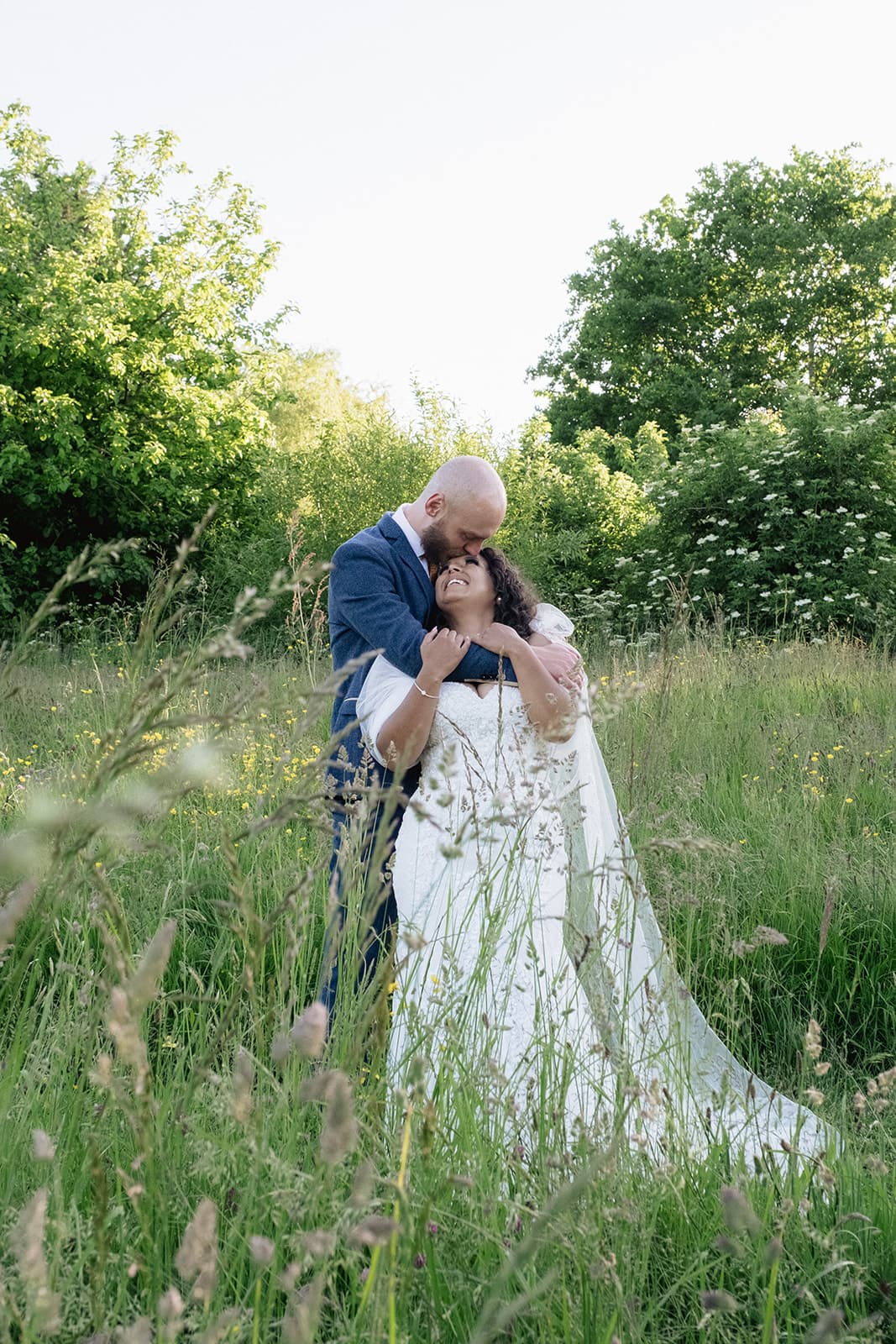 A bride and groom embracing in a sunlit meadow with trees in the background.
