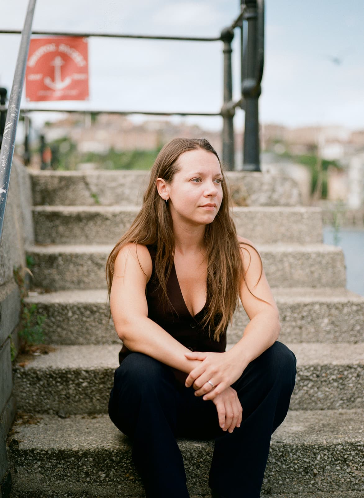 A woman with long brown hair sits on stone steps near the water, looking thoughtfully into the distance. A red sign with an anchor symbol is behind her. Photographed by Ivy & Pine