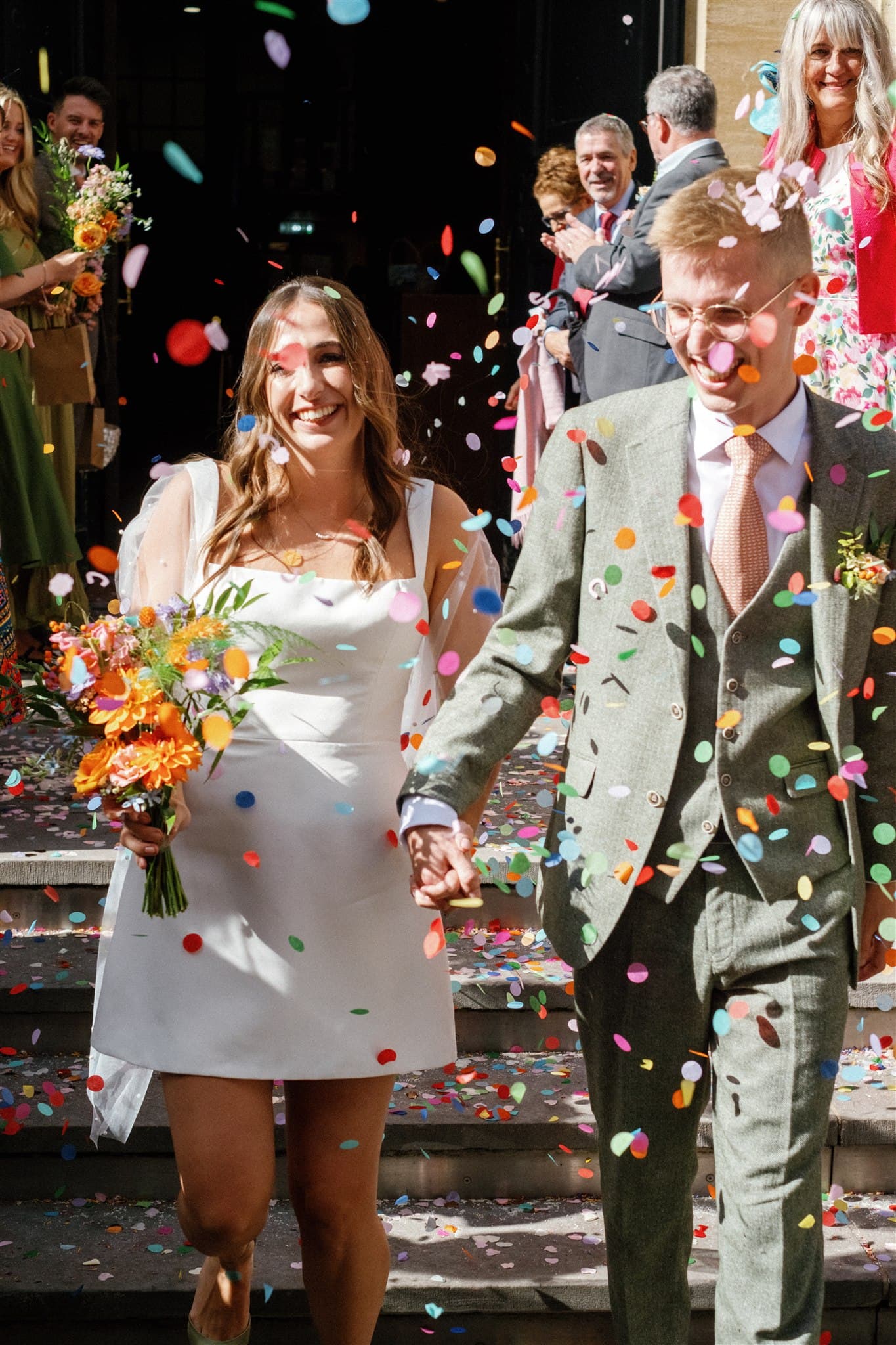 Bride and groom, smiling and holding hands, walk through colorful confetti thrown by guests. She holds orange flowers, and he wears a green suit.