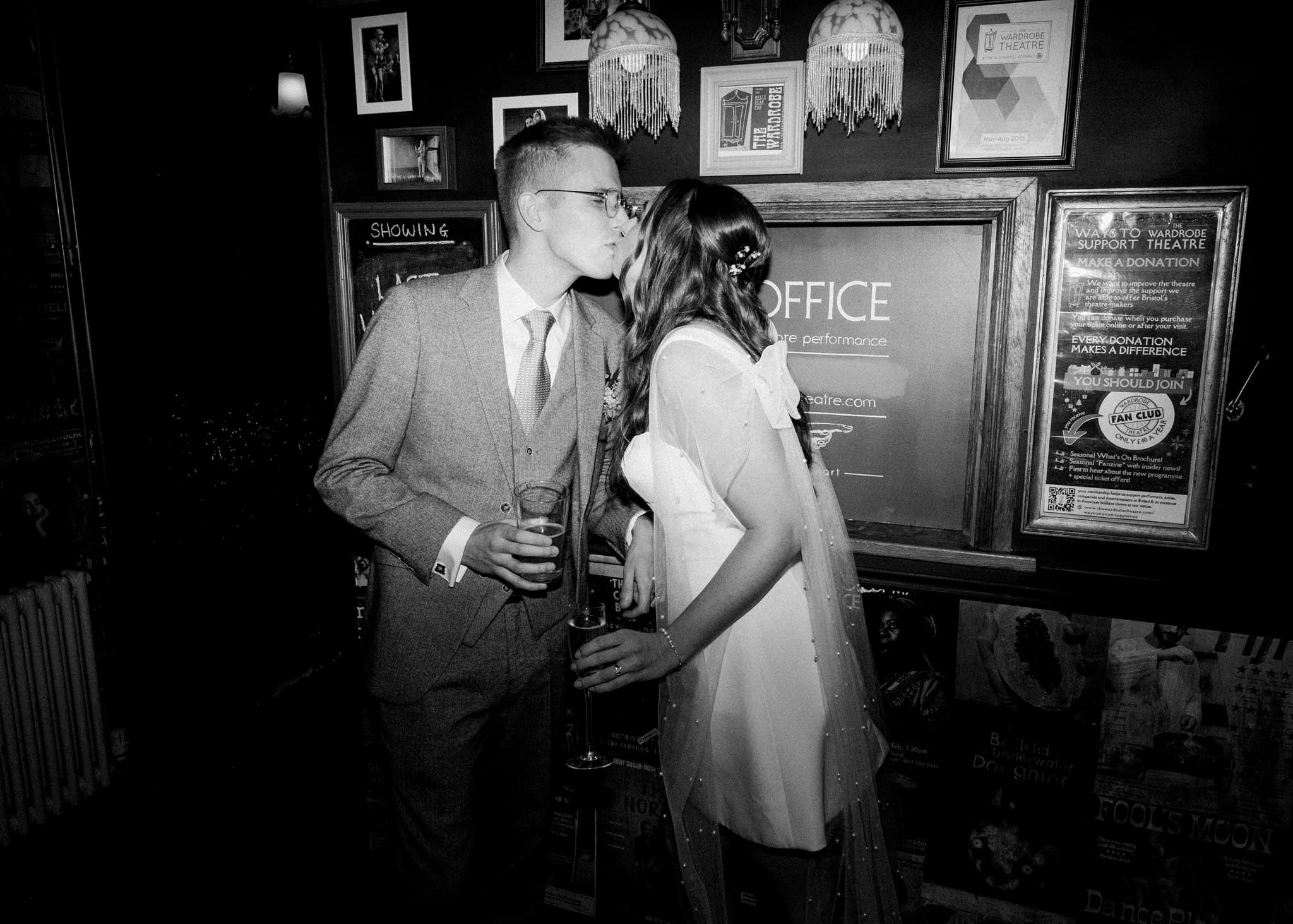 A couple kisses affectionately in a dimly lit, framed picture-filled hallway. The man wears a suit and glasses, and the woman a white dress, holding drinks.