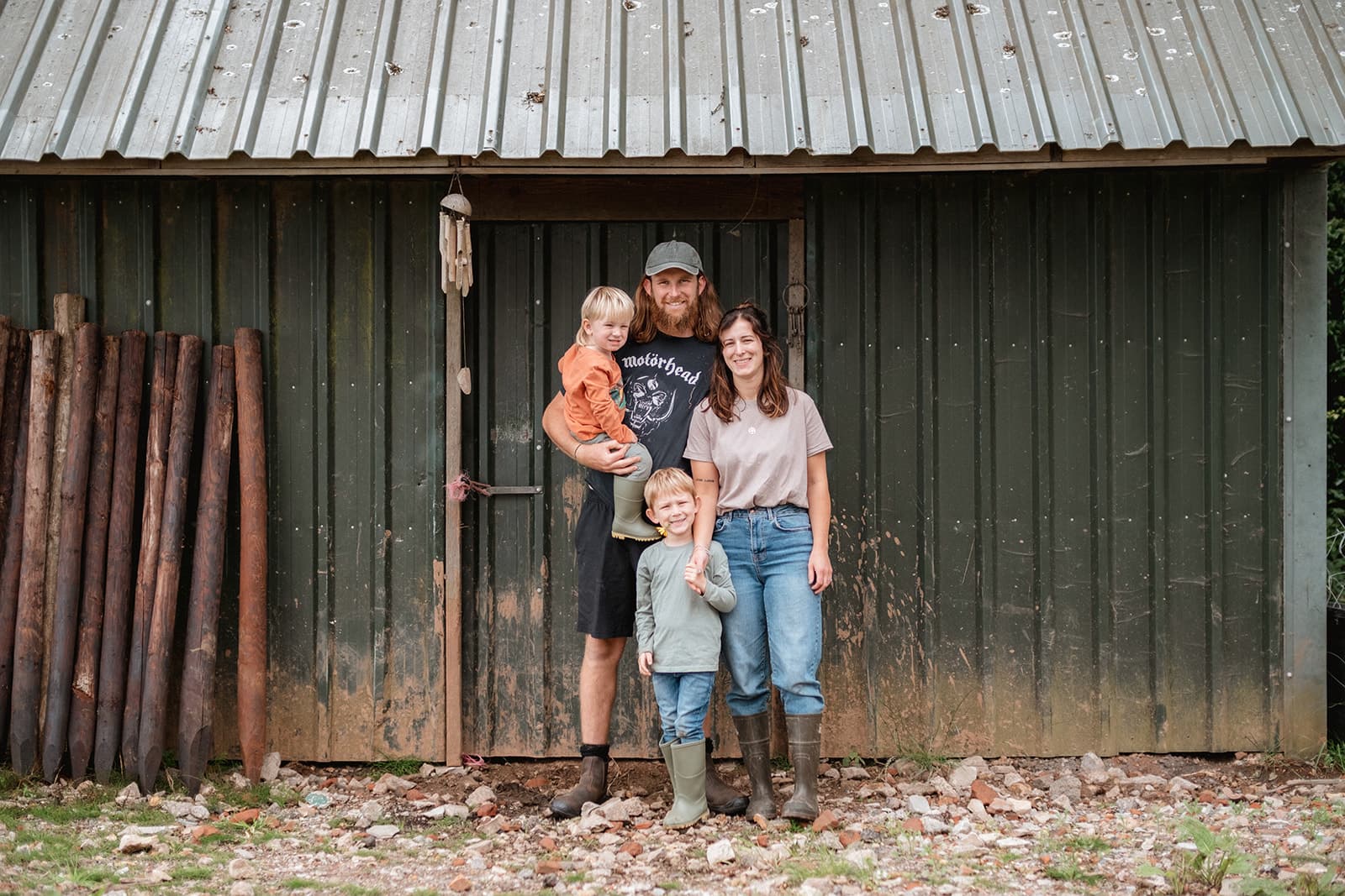 Three people in front of a rustic shed with corrugated roof and wooden logs stacked to the side.