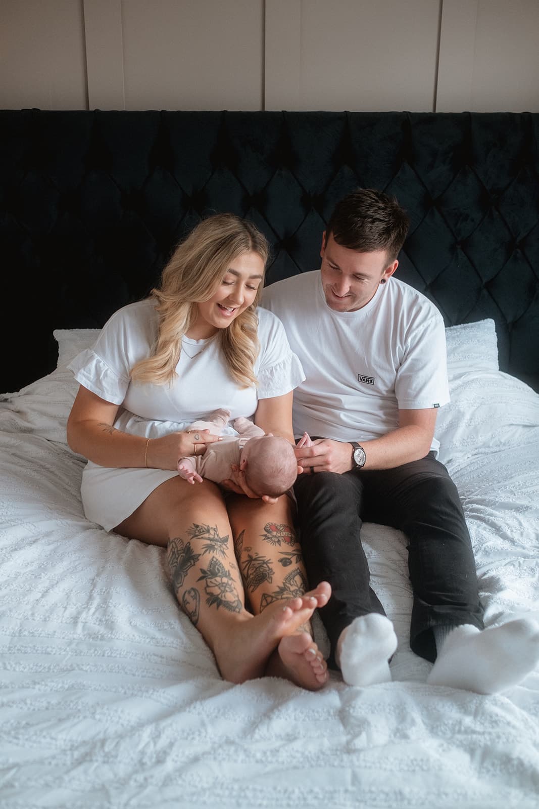 A couple sitting on a bed holding a baby, with tattooed legs visible.