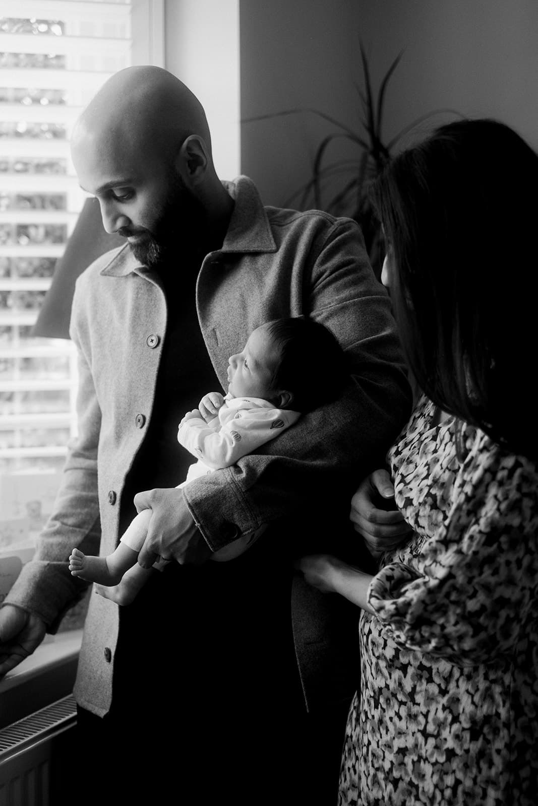 A black and white photo of two adults holding a baby, standing near a window.