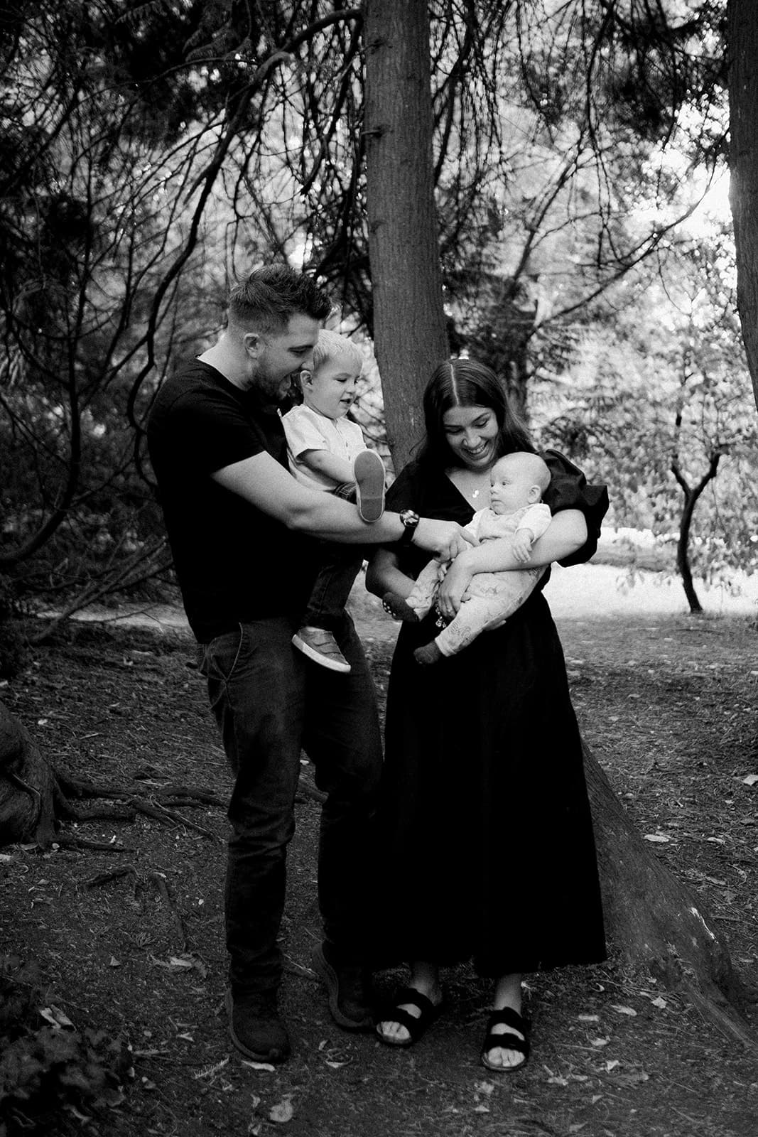 Two adults holding babies, standing in a wooded area.