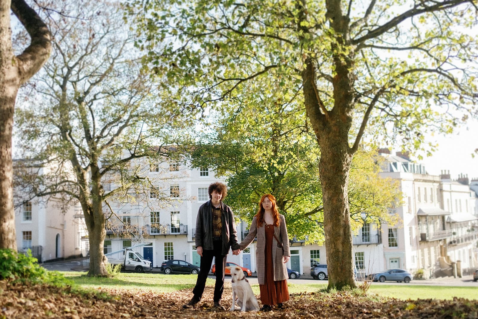 Two individuals and a dog standing under trees with autumn leaves, near white buildings.