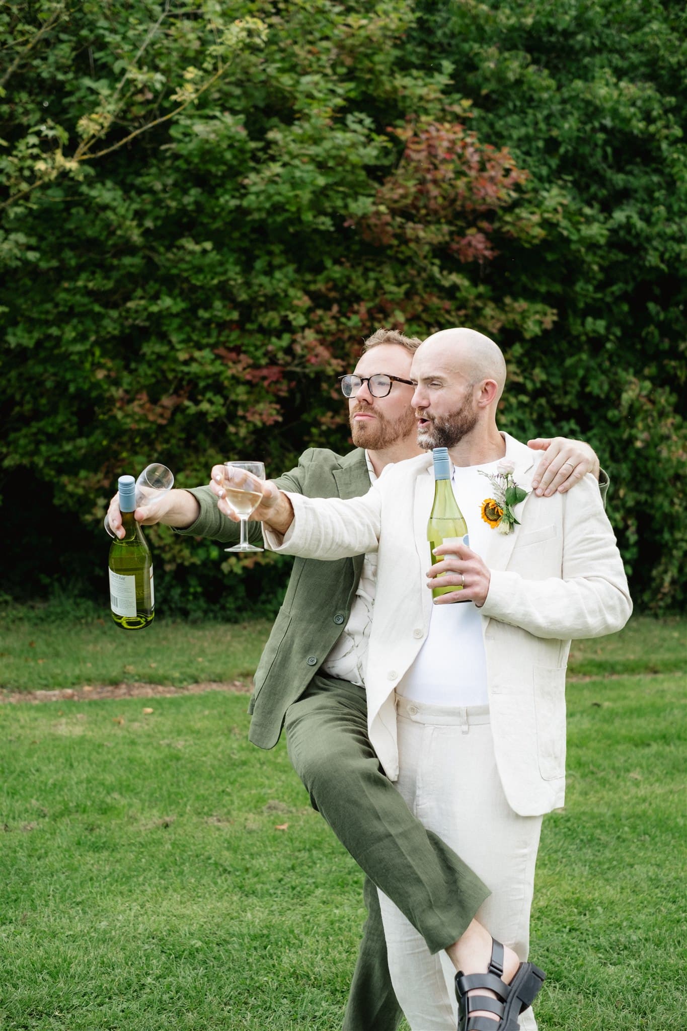 Person in a cream suit holding wine bottle and glass, with greenery in the background.