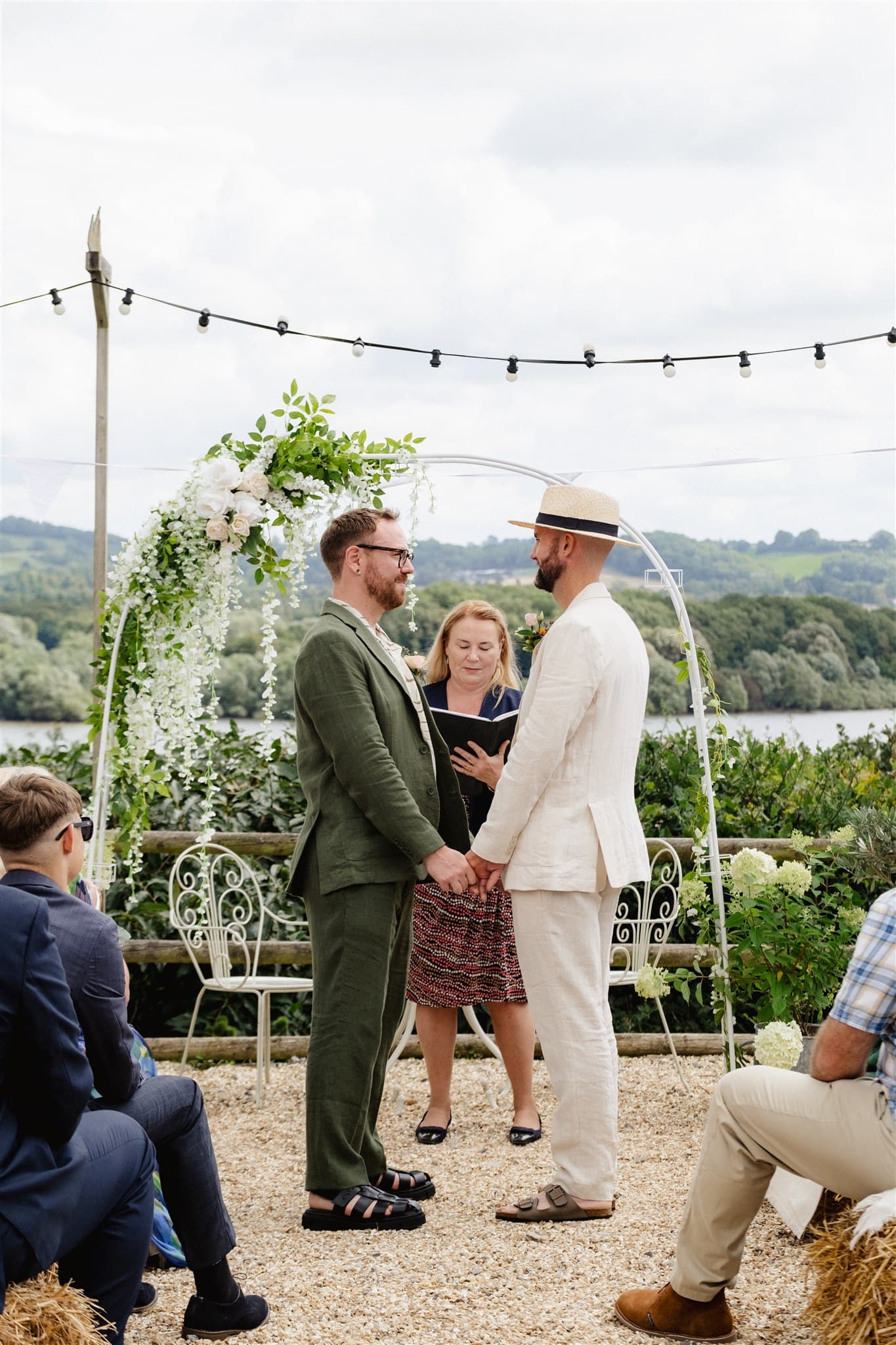 Outdoor wedding ceremony with a floral arch and guests seated on hay bales.