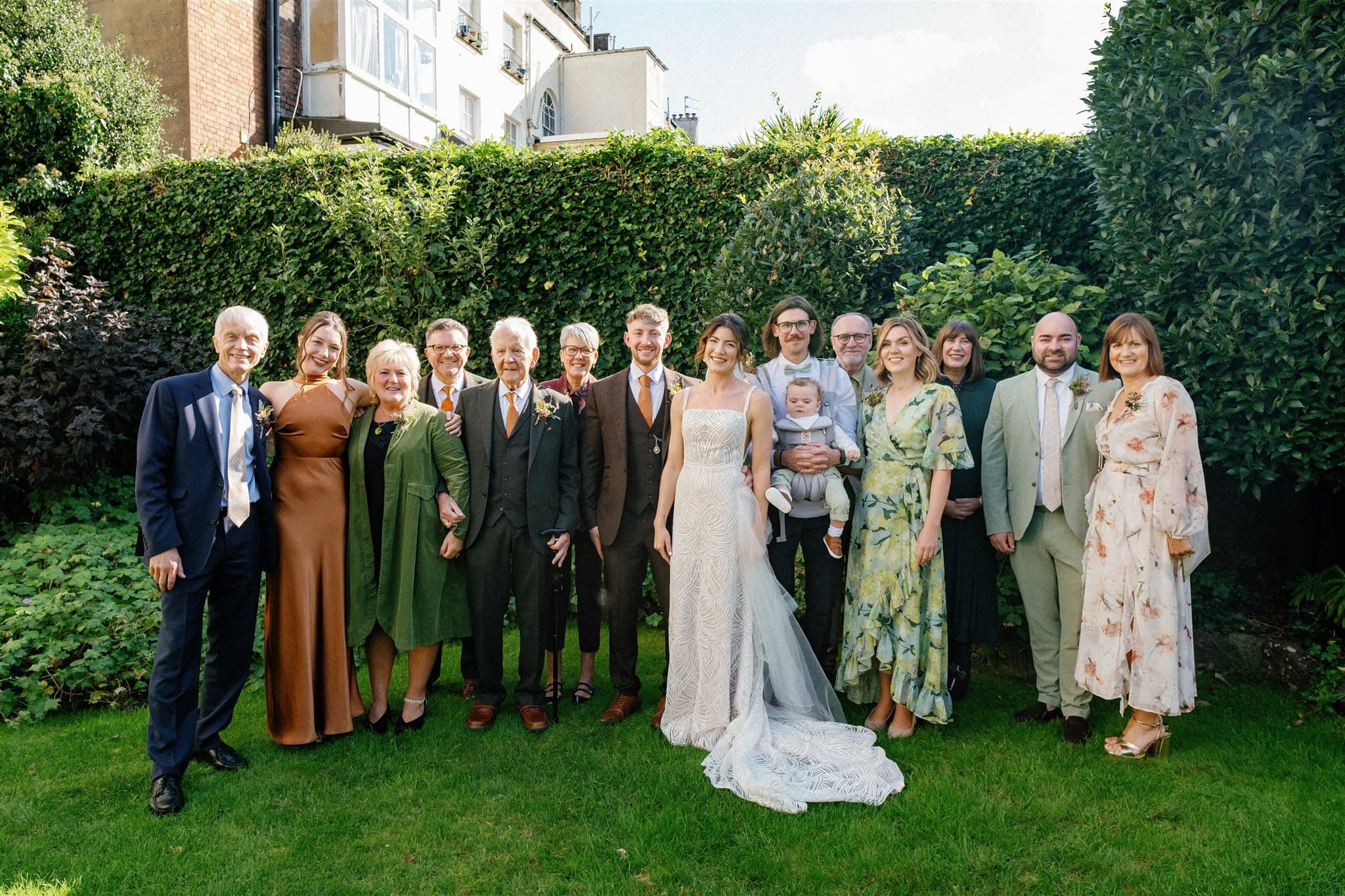 A group of elegantly dressed people at an outdoor event, standing on grass with a hedge backdrop.
