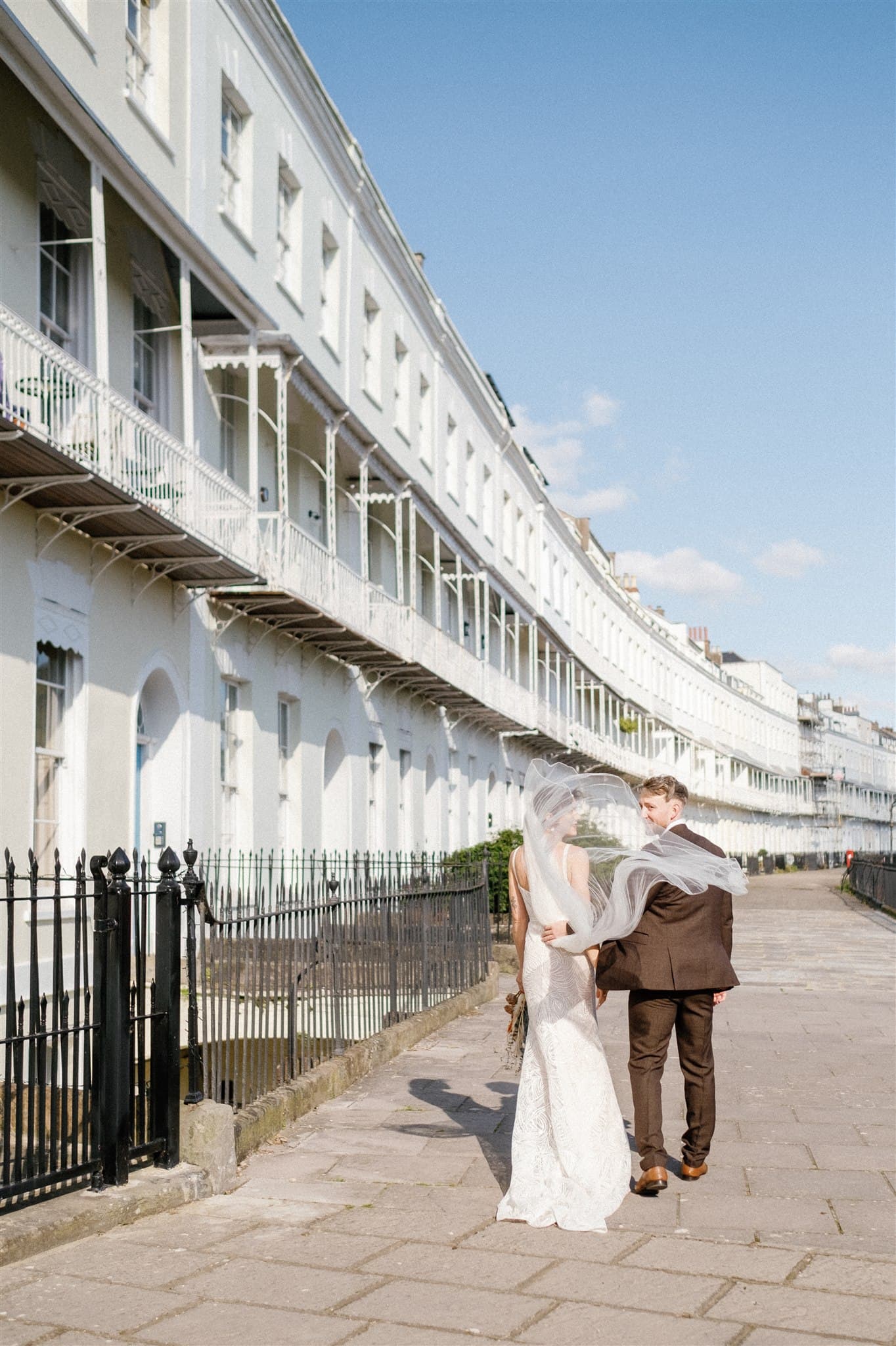 Bride and groom walking hand in hand by a row of houses.