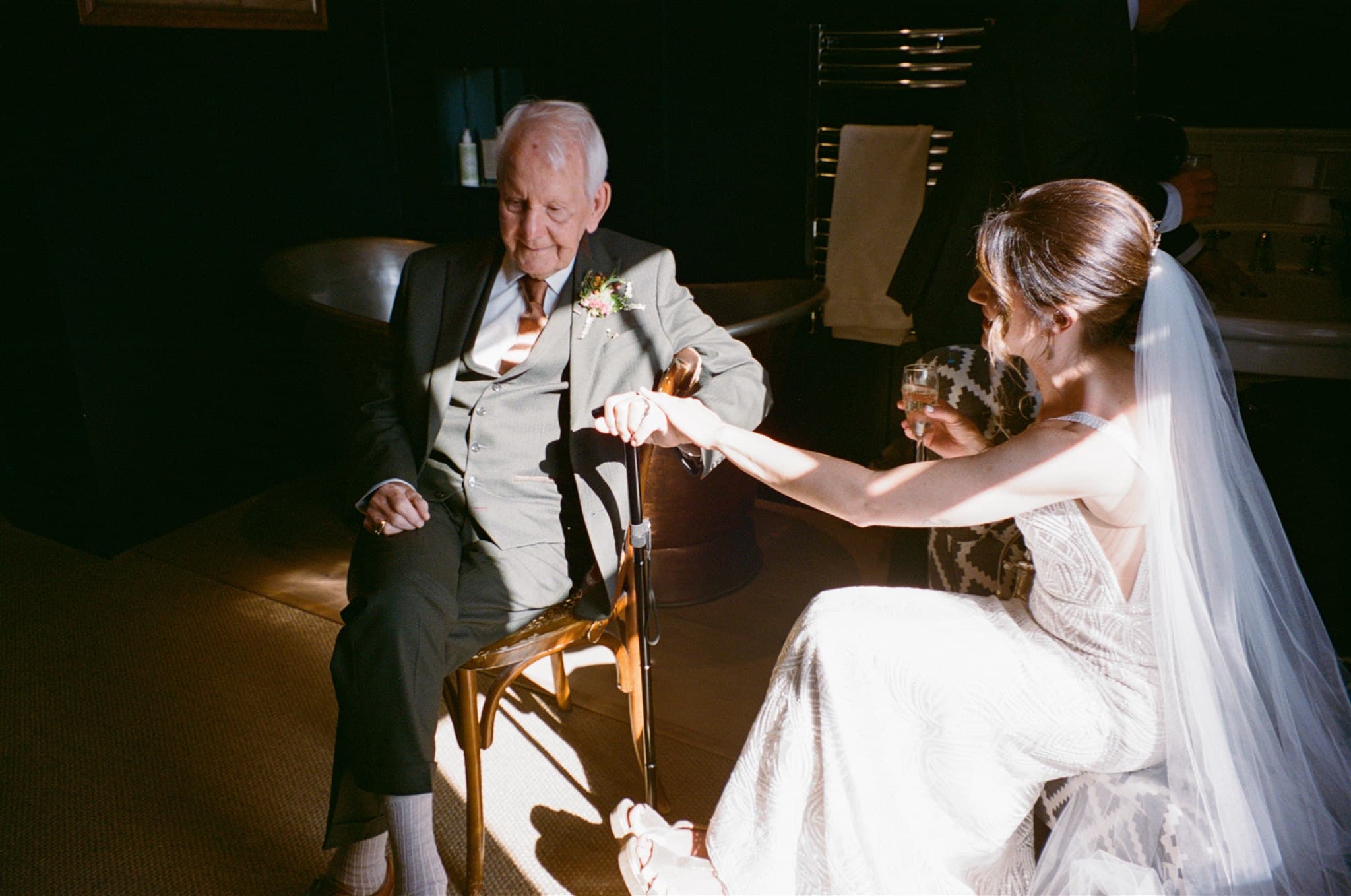 Bride in white dress holding a drink, sitting opposite a suited individual with a cane.