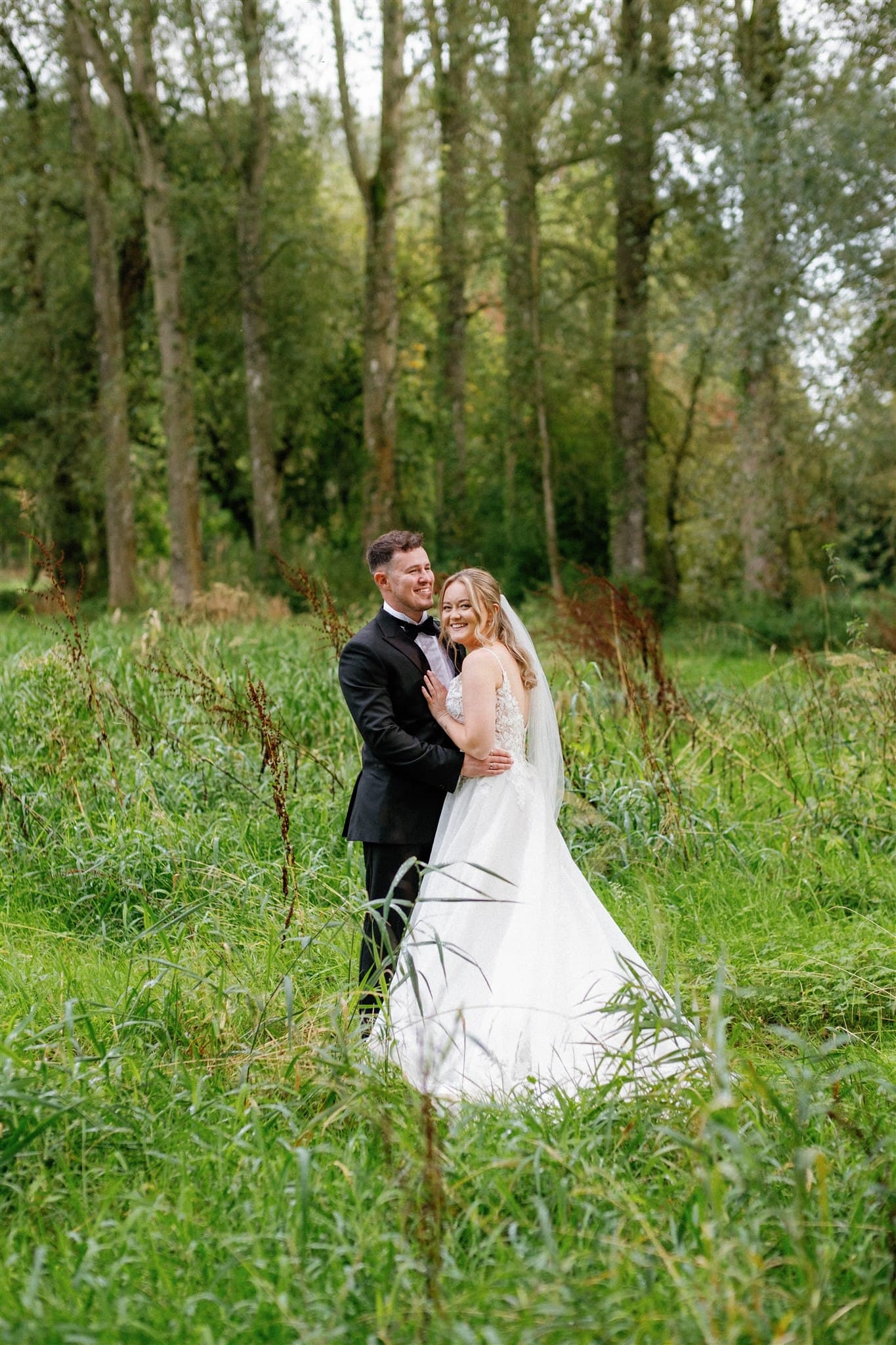 A bride and groom embracing in a lush green field with trees in the background.